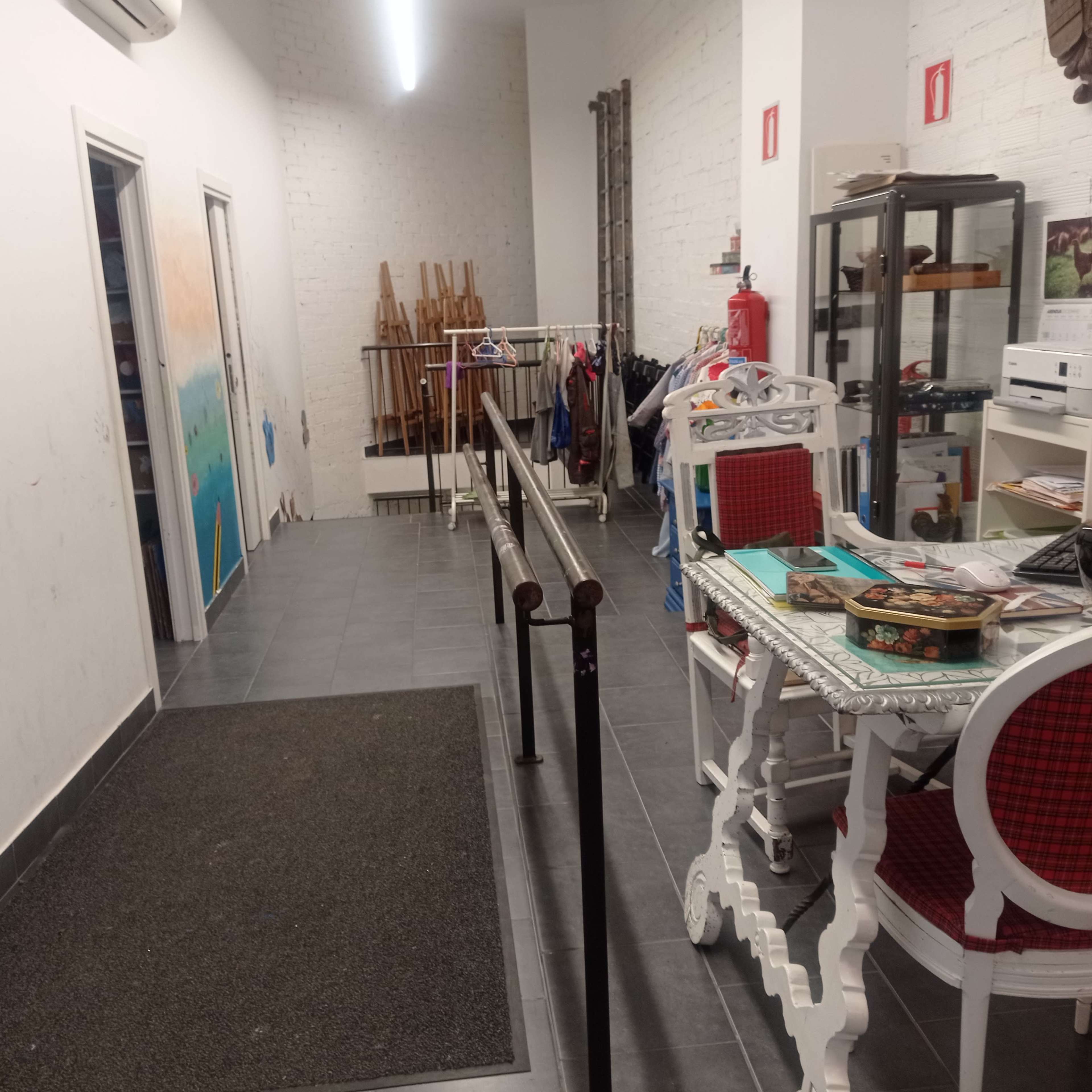 The image shows a hallway with clothing racks on one side, a table with various items on the other, and doors leading to different rooms.