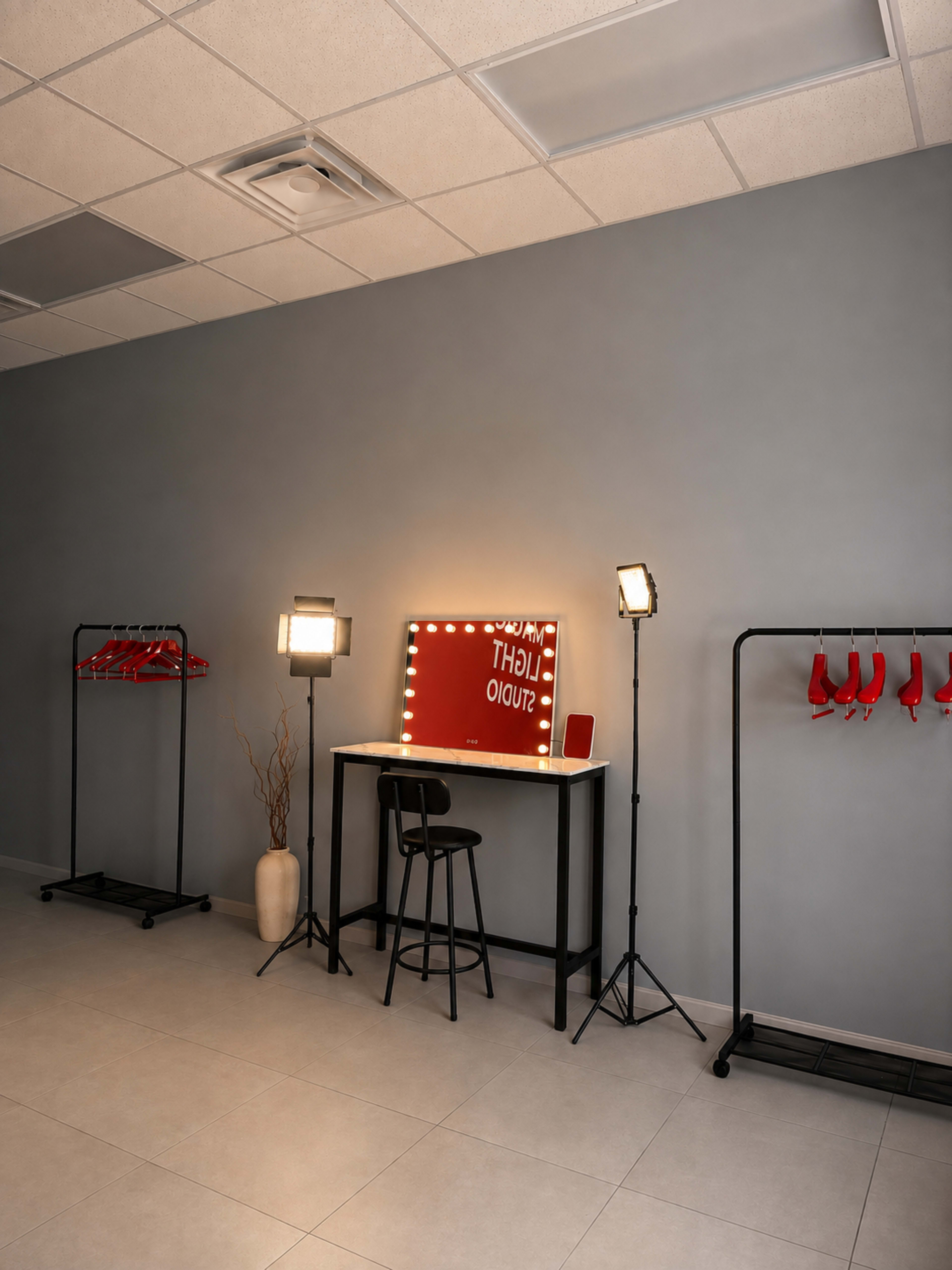 The image shows a neatly arranged studio space with a mirror surrounded by lights, a black stool, and racks displaying red clothing.