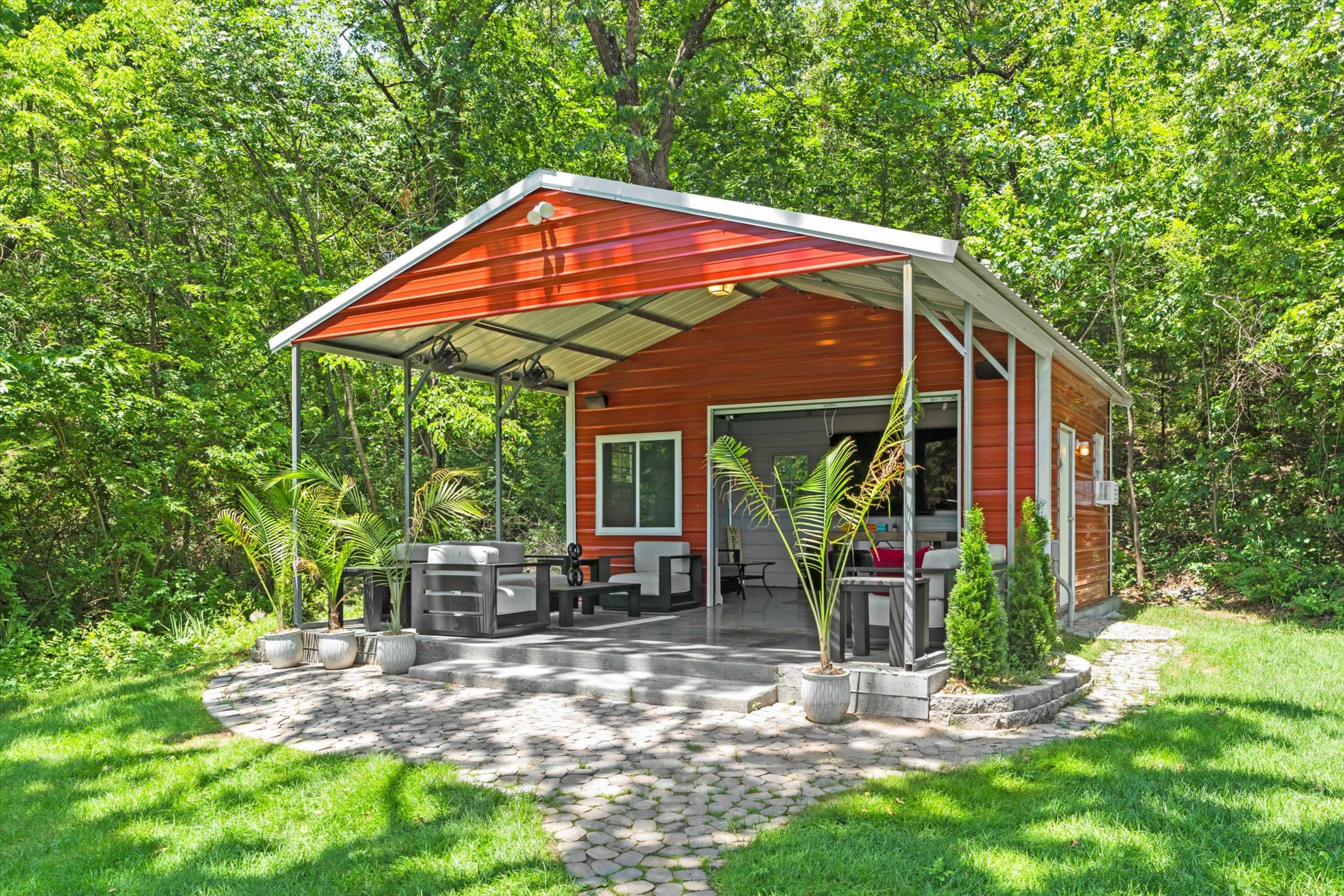 A small cabin with a wooden exterior and a metal roof sits on a stone patio surrounded by green vegetation.