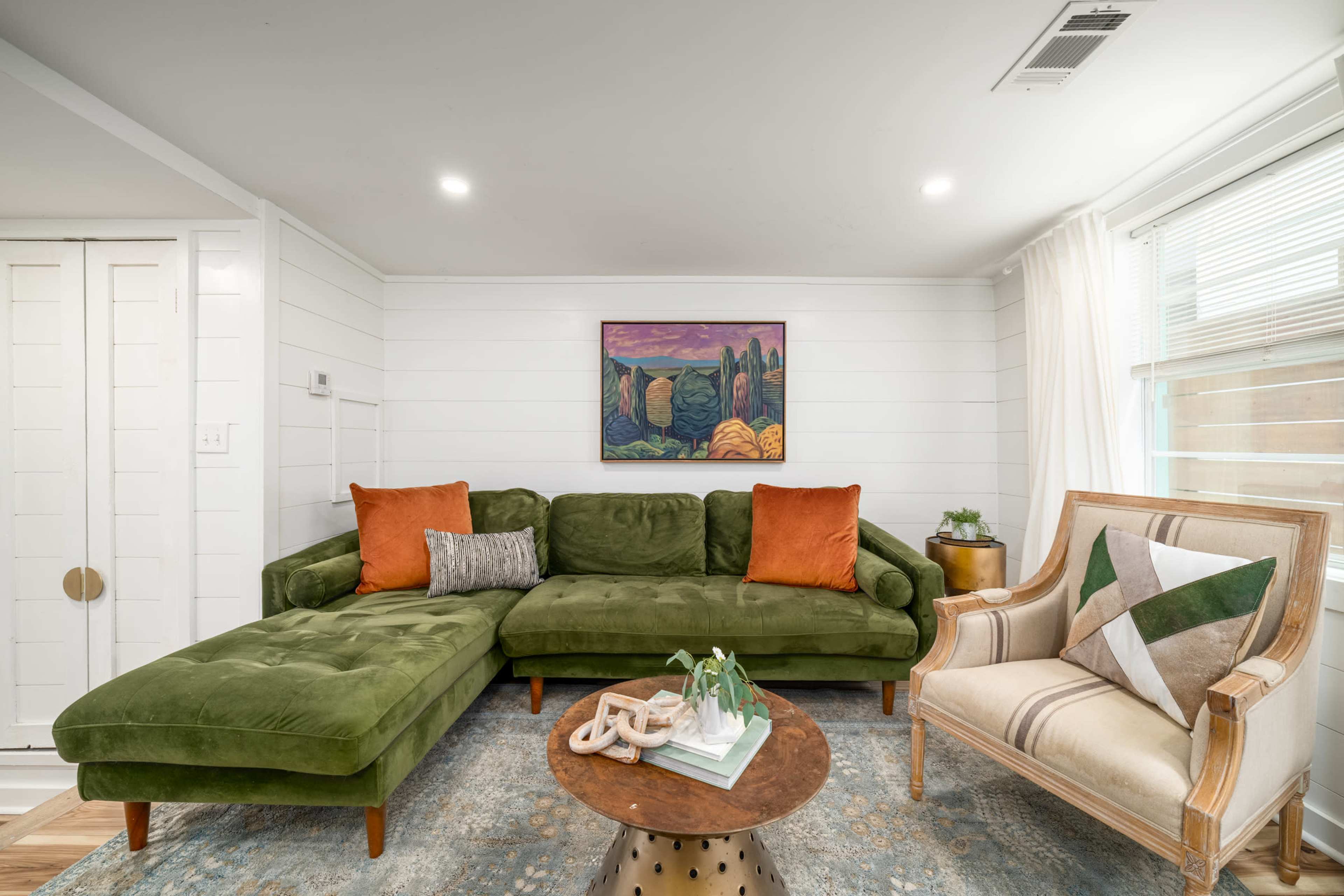 The scene features a cozy living room with a green sectional couch, two orange throw pillows, an accent chair, a round coffee table, and a colorful painting on the wall.