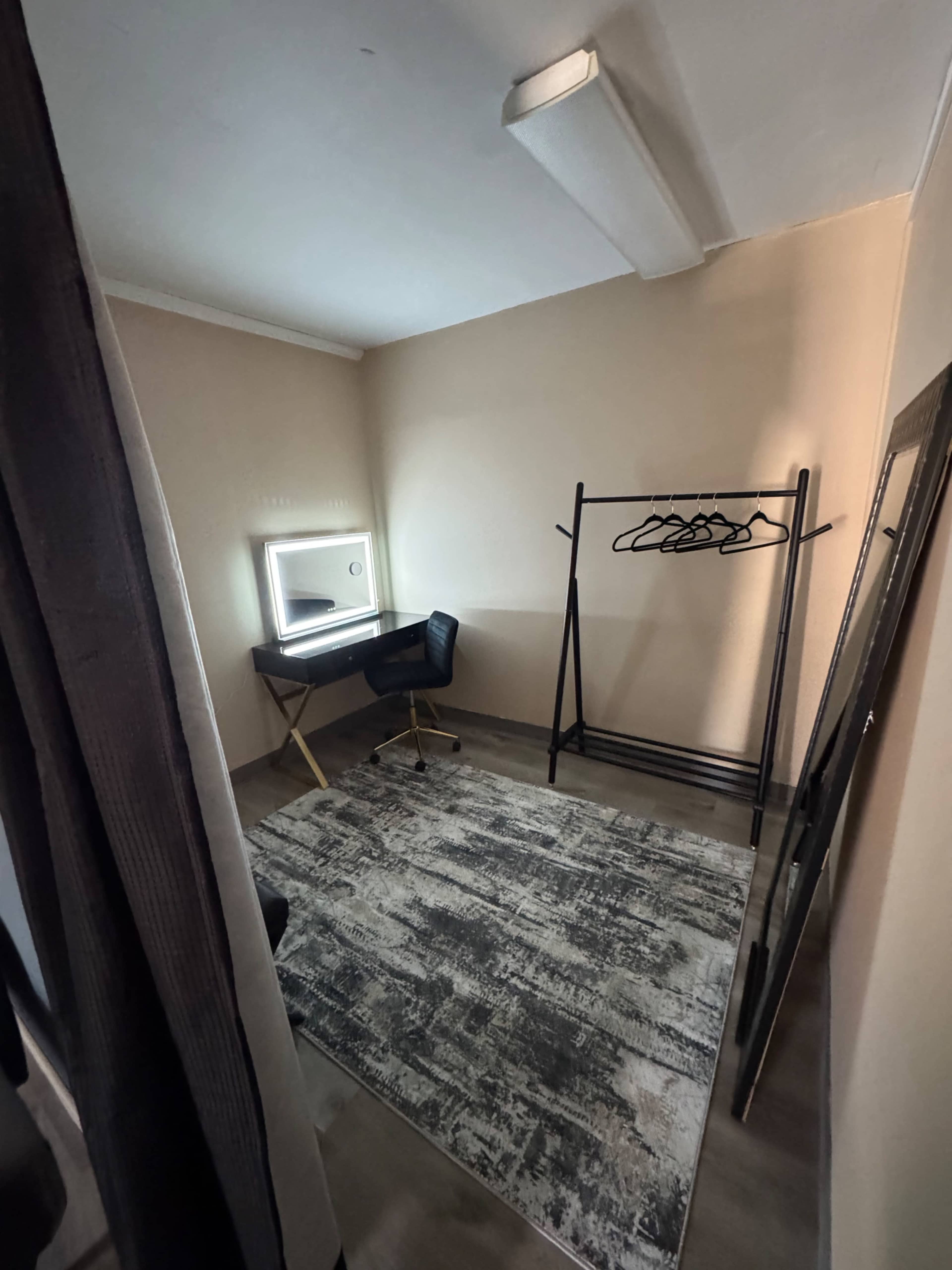 The image shows a small, minimally furnished room featuring a vanity with a lighted mirror, a black chair, a clothing rack, and a large mirror against a beige wall.