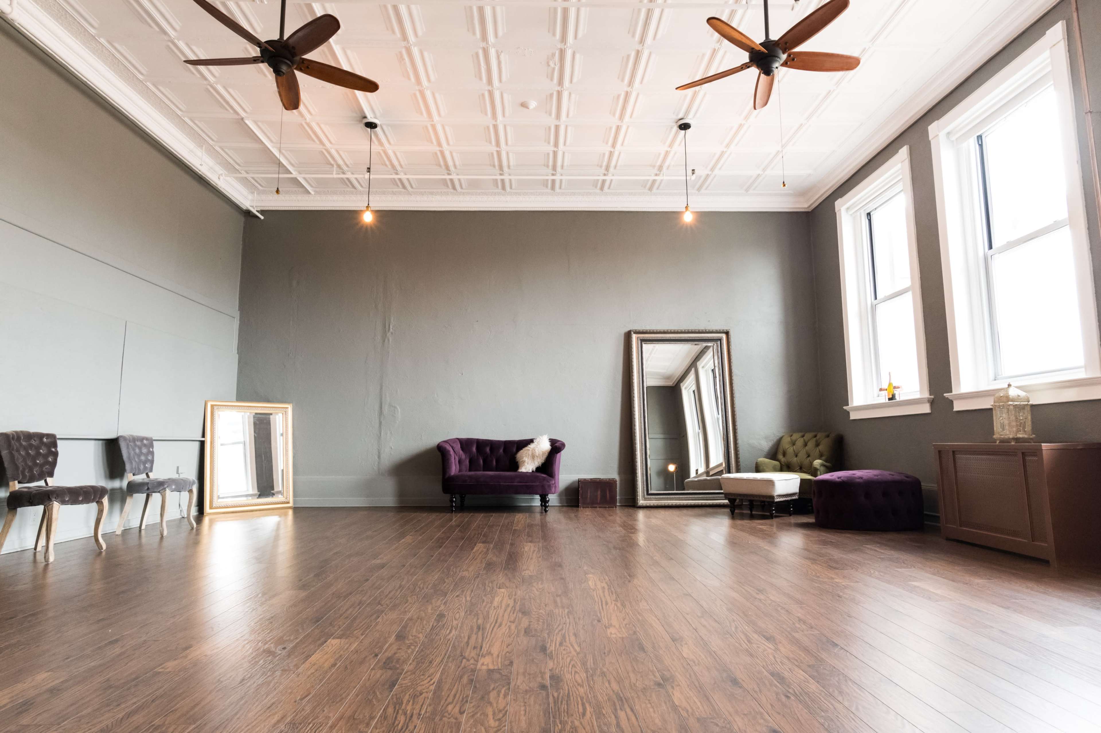 A spacious room with wooden flooring, two ceiling fans, large windows, and a variety of furniture including a purple sofa, a white chaise lounge, and decorative mirrors.