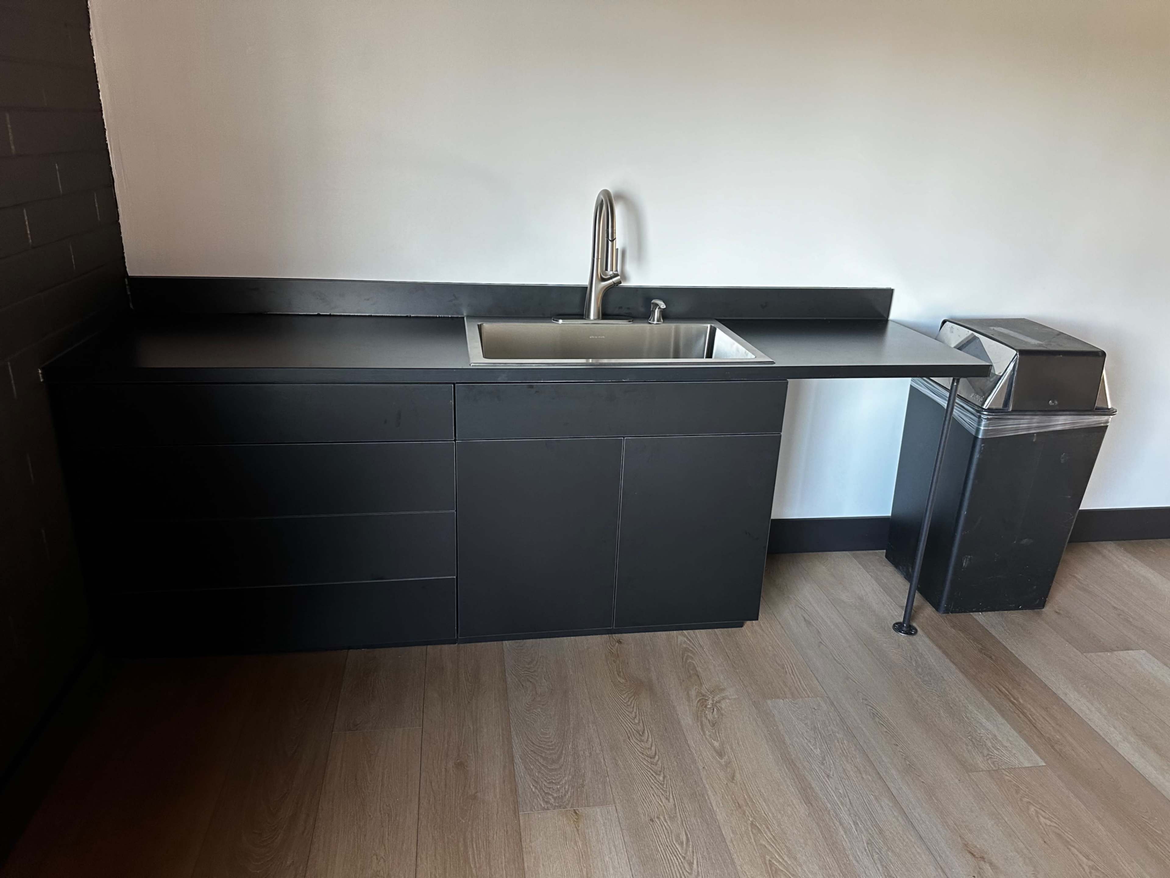 A modern kitchen area featuring a black sink and cabinetry alongside a gray trash bin on a wooden floor.