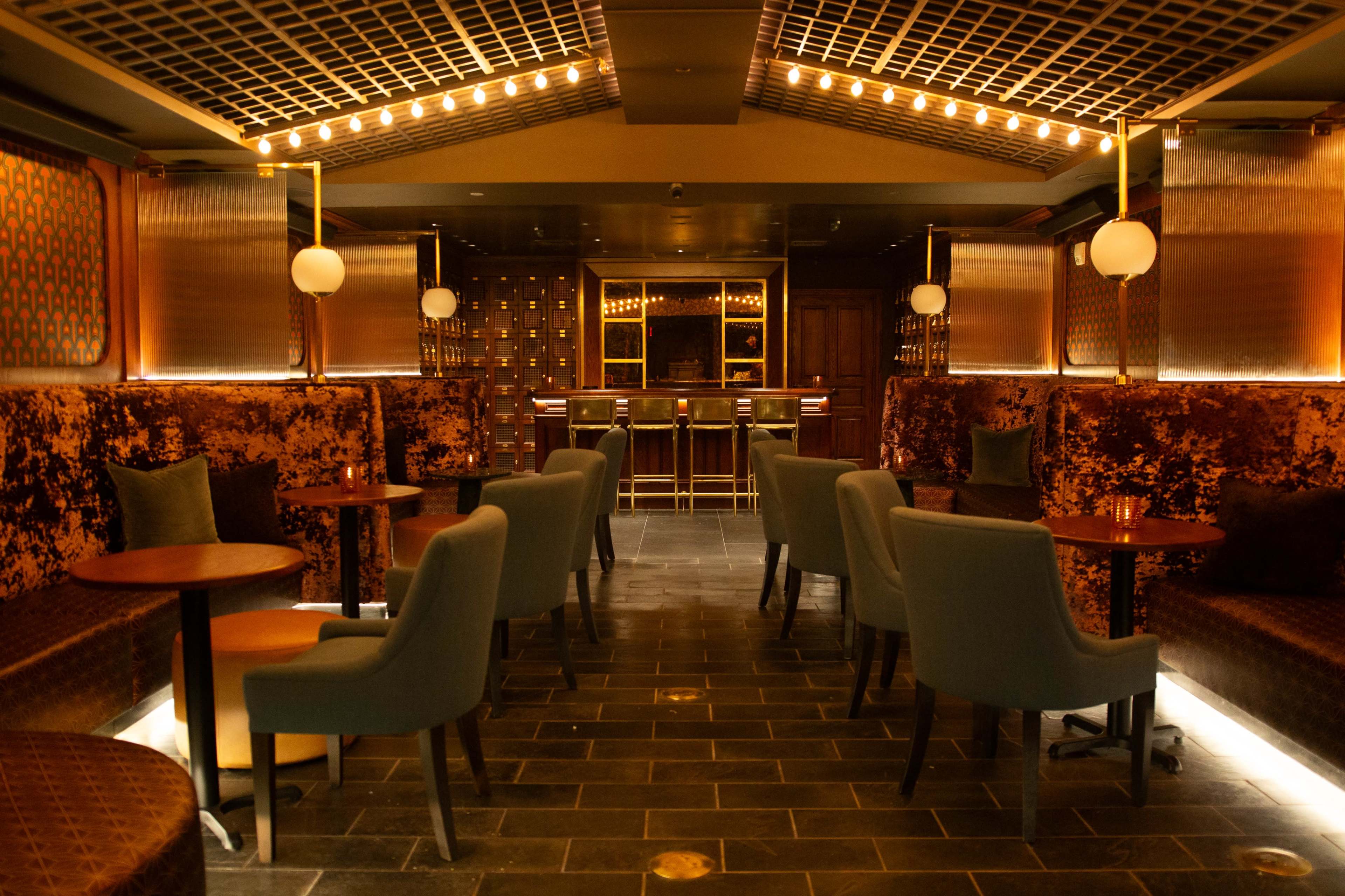 The image shows a dimly lit lounge with plush seating, small round tables, and a bar at the end of the room.