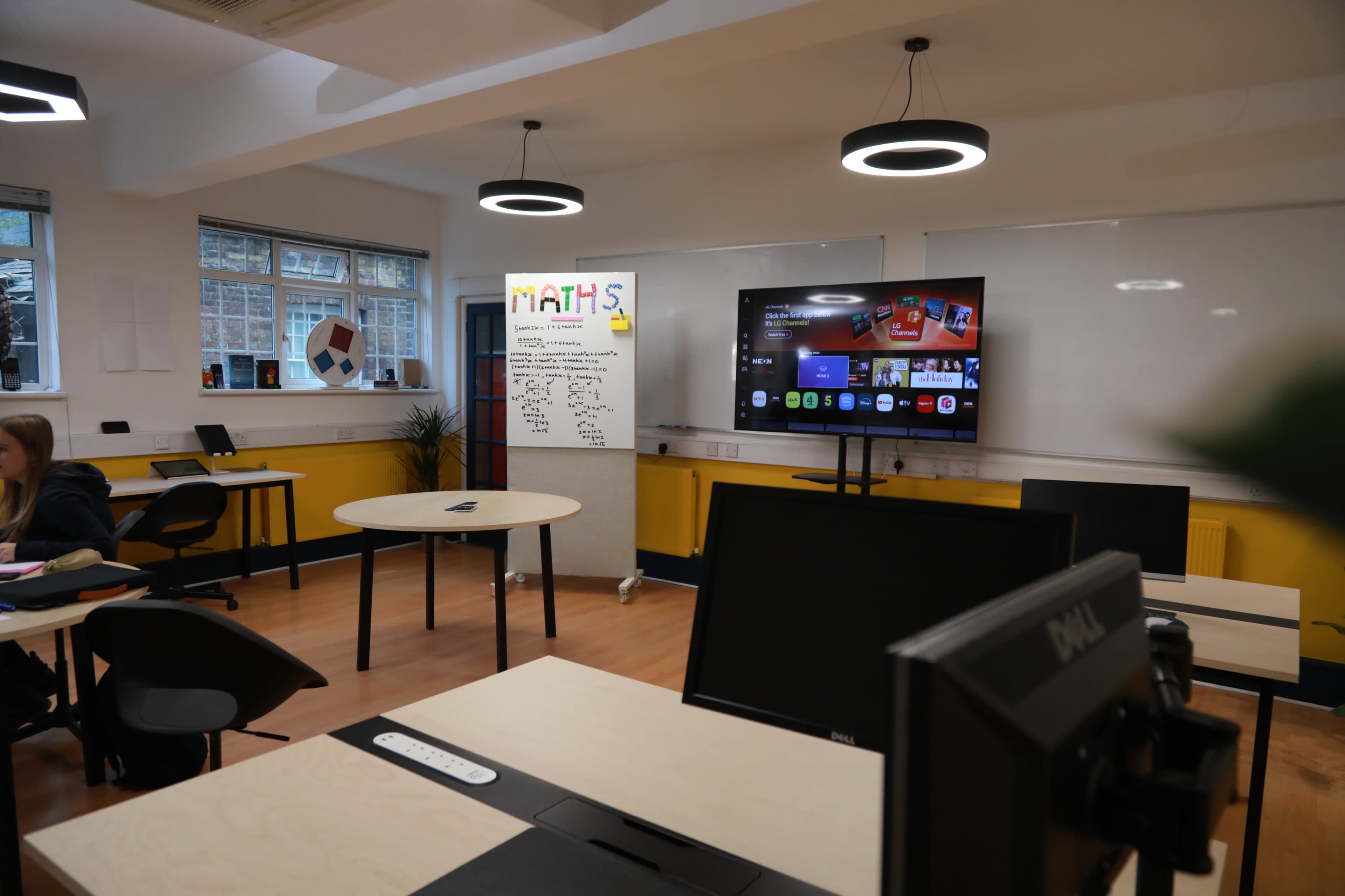 The image depicts a modern classroom featuring a large screen displaying educational content, with round tables and black chairs arranged around the space.