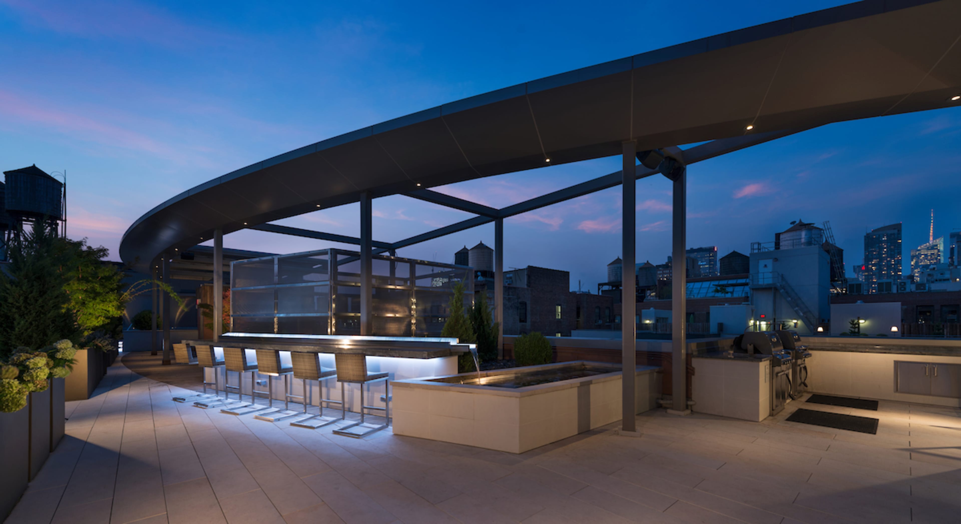 A modern rooftop terrace features a sleek bar area, planters, and a view of a city skyline at dusk.