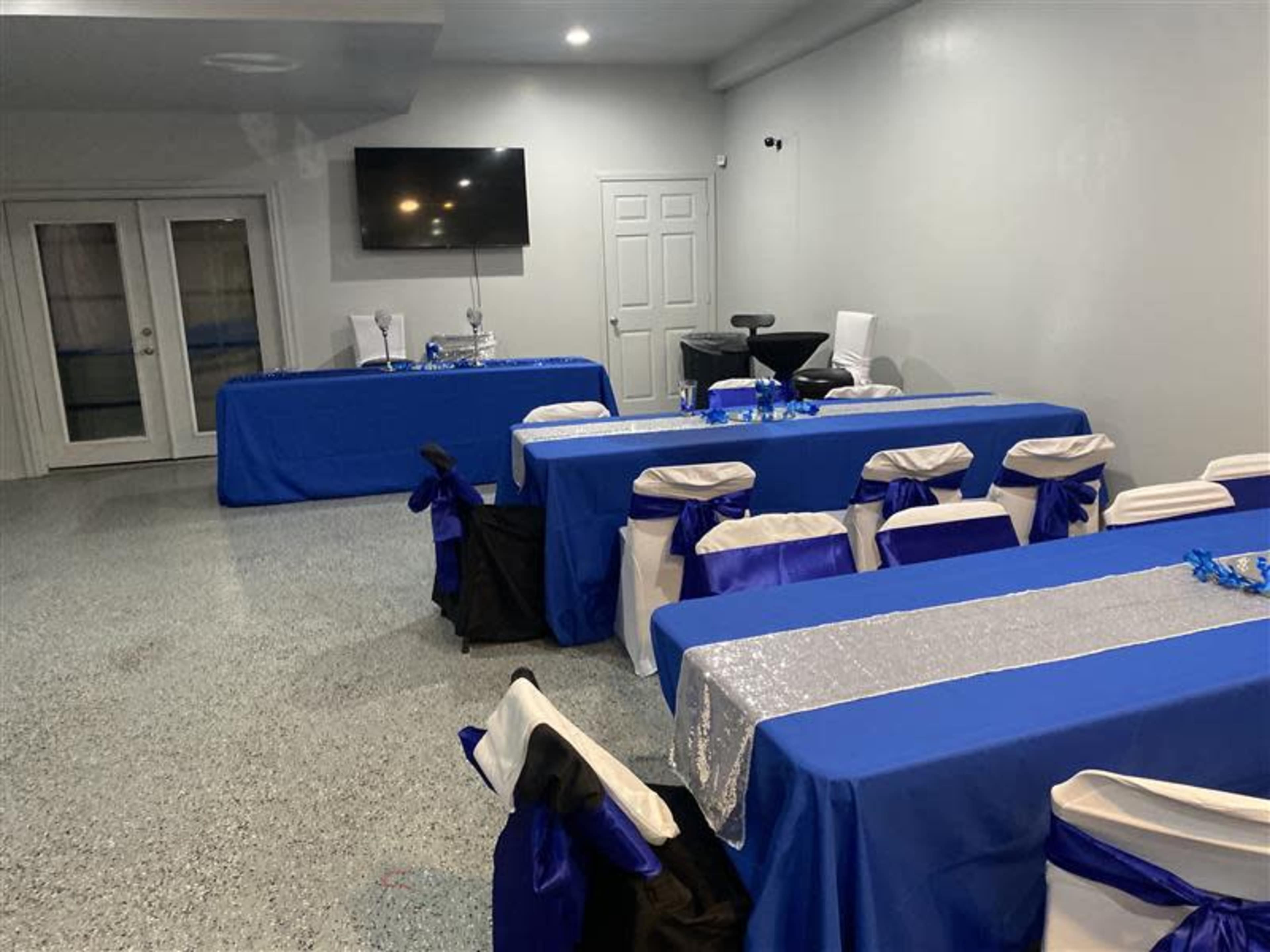 A room set up for an event, featuring tables covered with blue tablecloths and white chairs adorned with blue sashes, along with a TV and a side table.