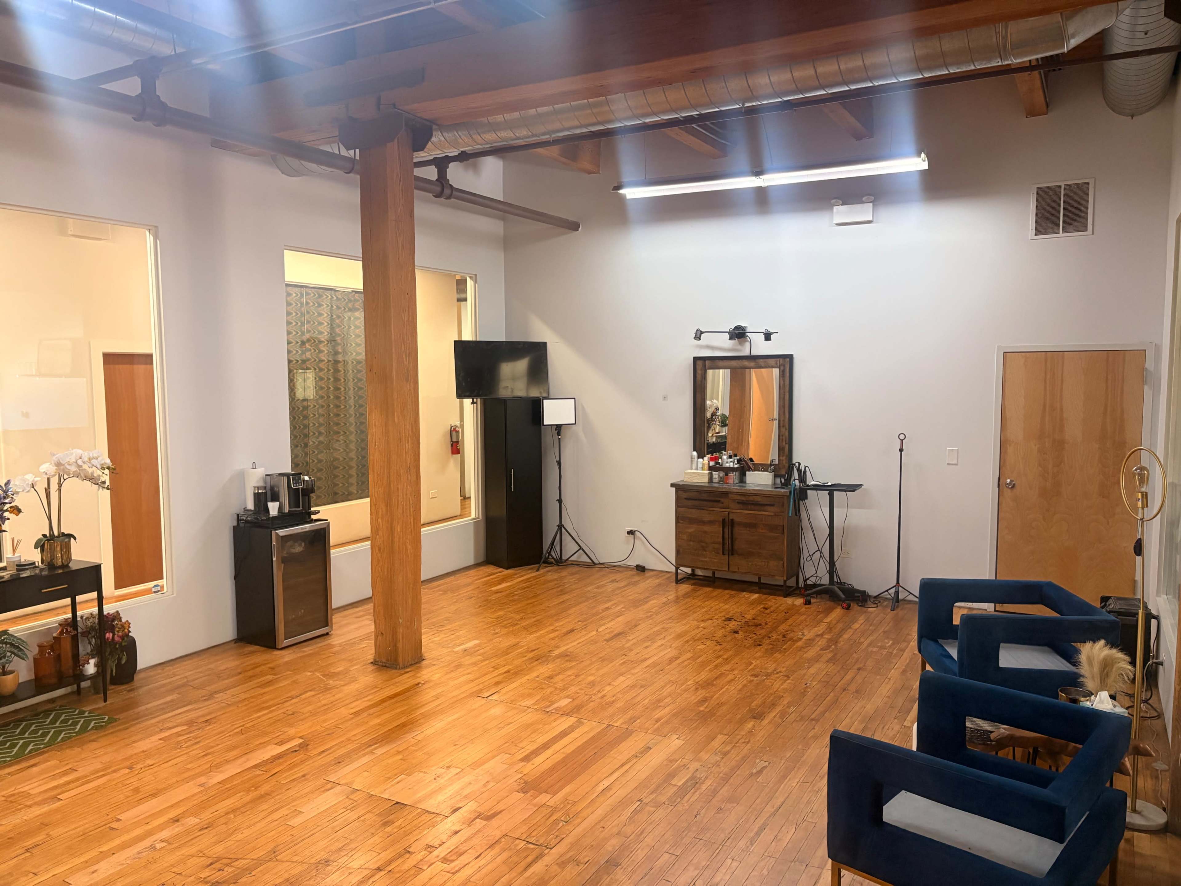 The image shows a spacious, well-lit room with wooden floors, a large mirror, a wooden cabinet, and blue chairs arranged around the area.