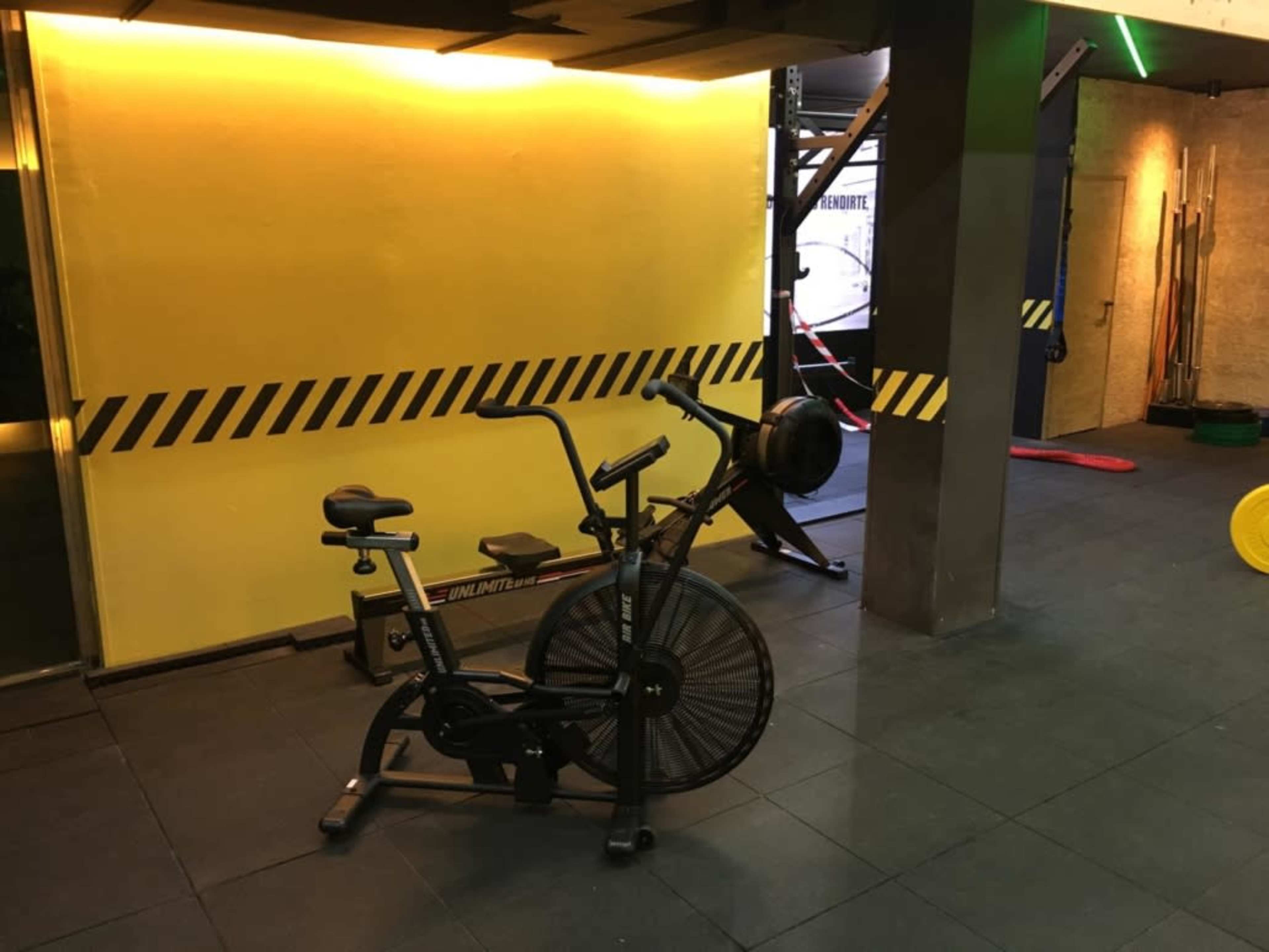 A fitness area with a stationary bike and a rowing machine against a yellow wall with black striped caution tape.
