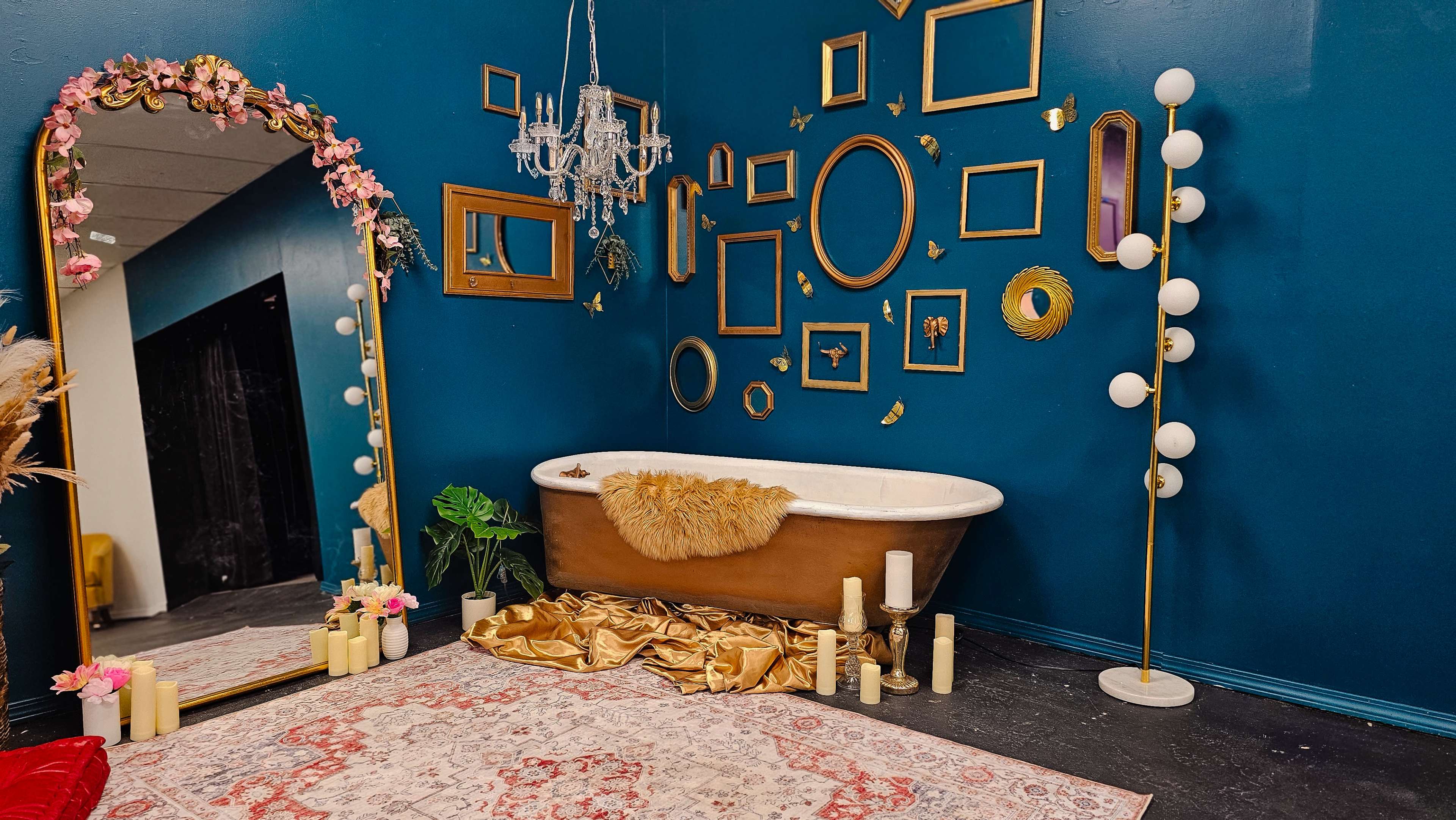 The scene features a vintage freestanding bathtub near a vibrant blue wall adorned with various empty picture frames, a chandelier above, and decorative elements like flowers and candles on the floor.
