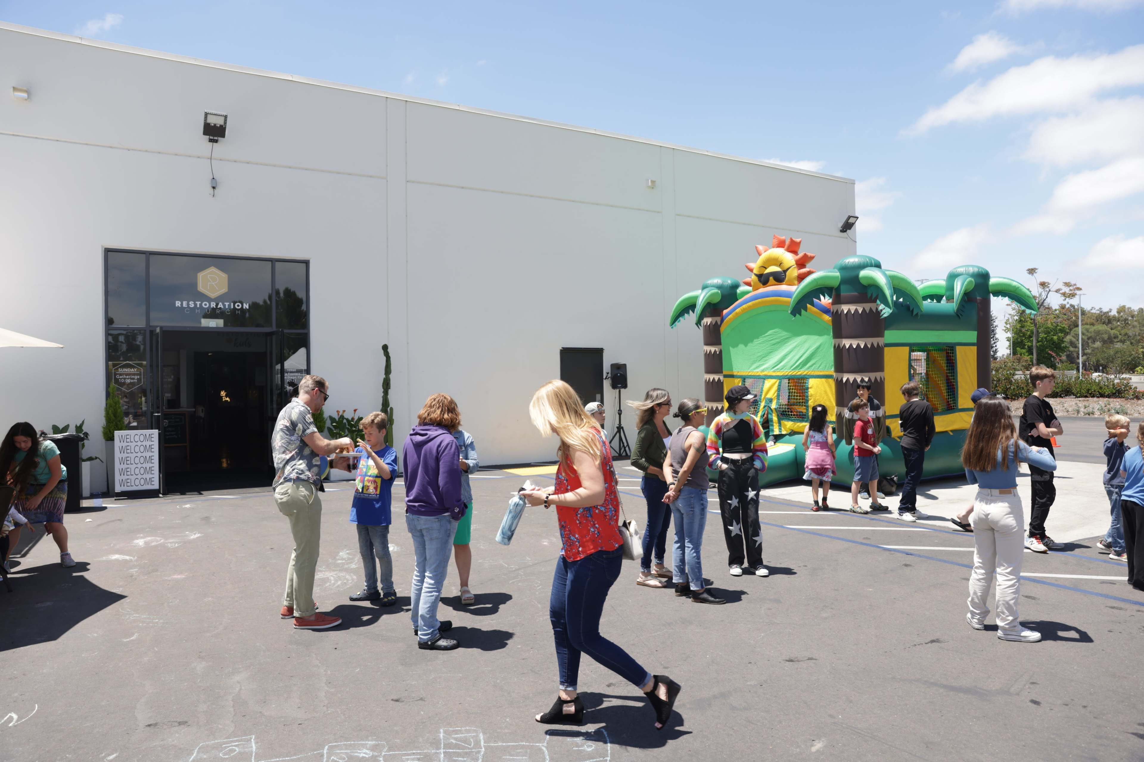 A crowd gathers outside a building with a sign reading "Restoration" while a colorful inflatable bounce house shaped like palm trees is set up nearby.