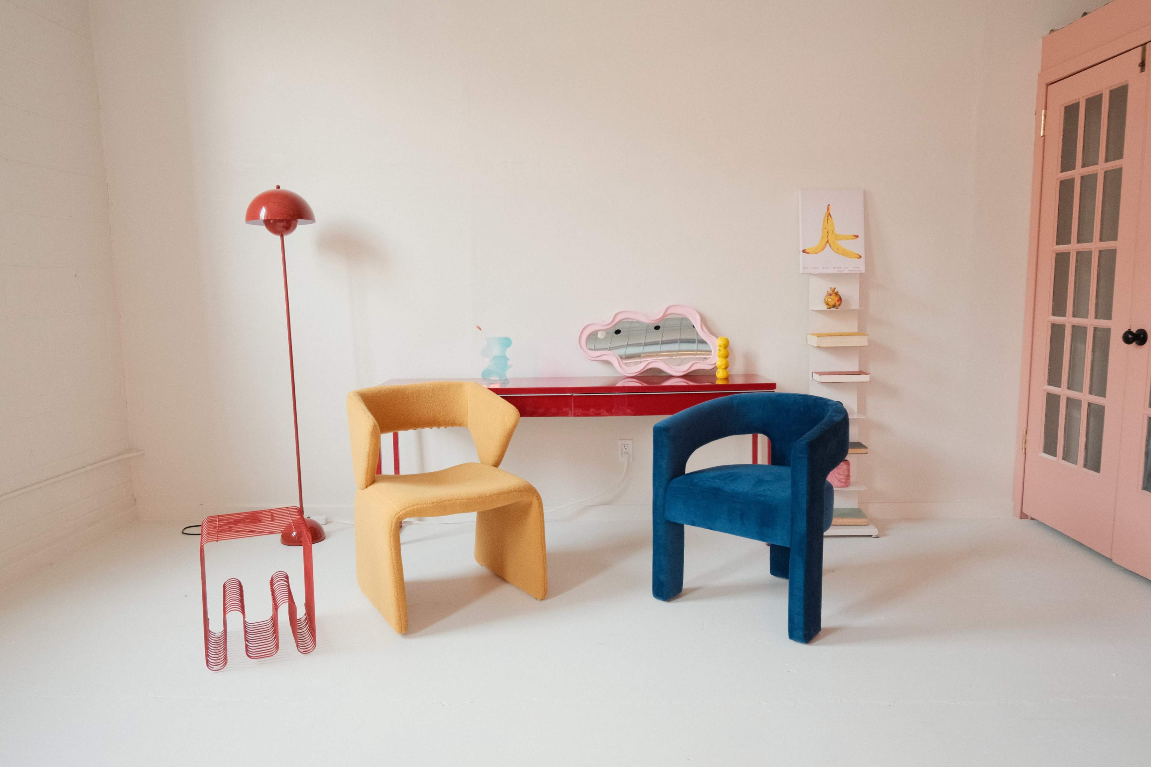 The image features a minimalist room with a yellow and a blue chair, a red lamp, a red desk, and decorative shelves against a white wall.