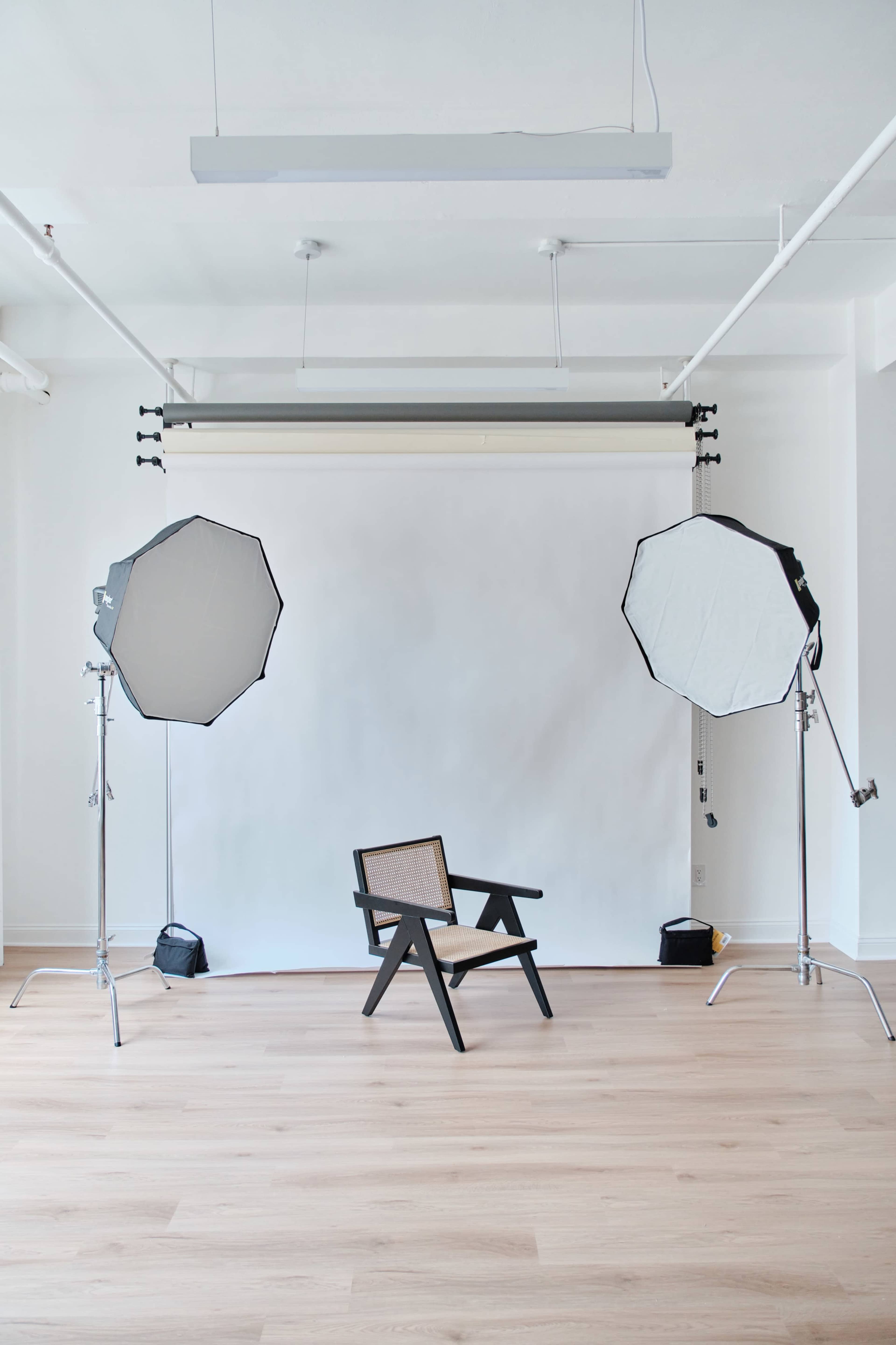 A minimalist photography studio features a black chair positioned in front of a white backdrop, flanked by two large softbox lights.