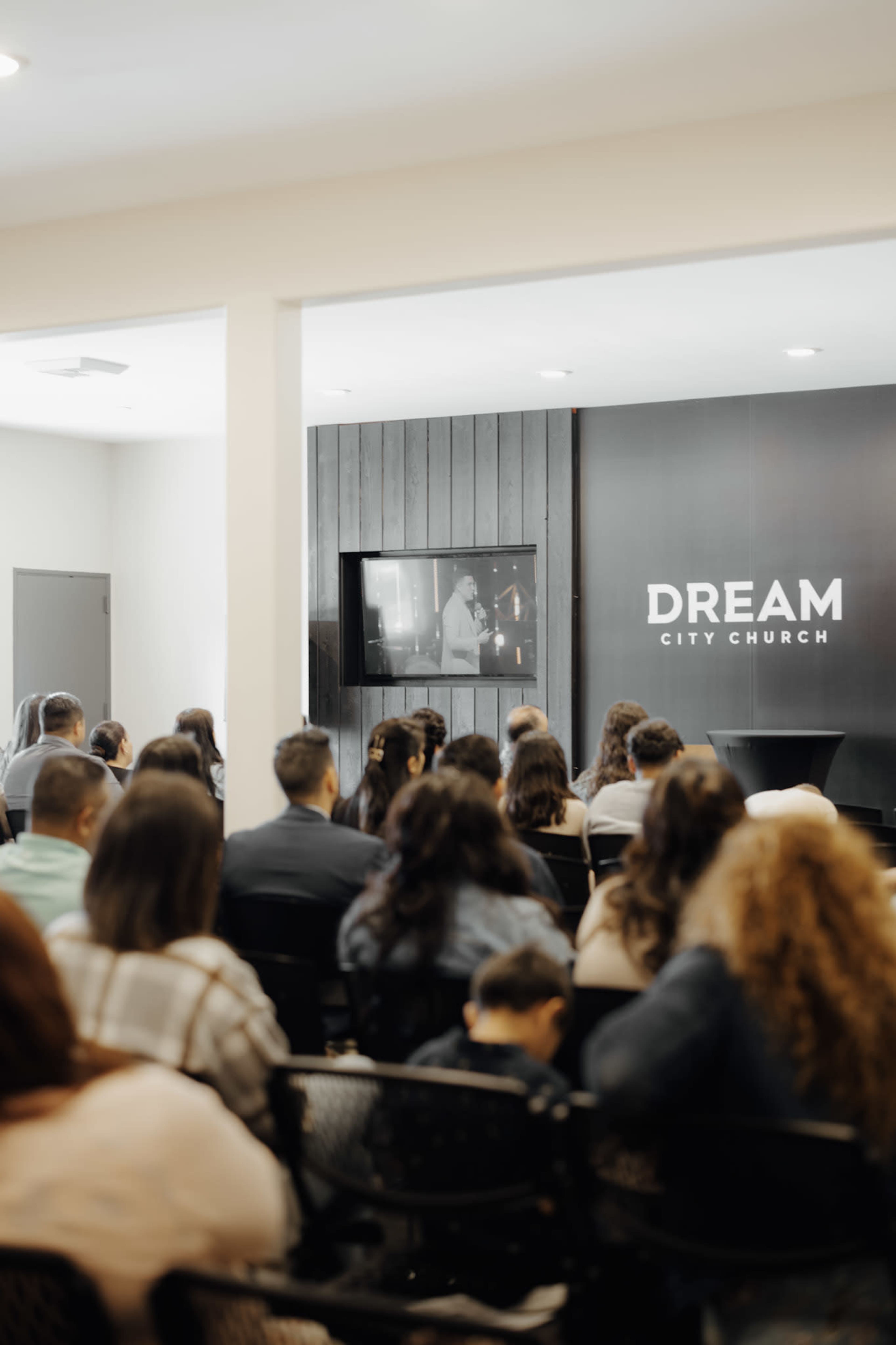 A congregation sits in rows of chairs, facing a screen displaying a presentation in a modern church setting.