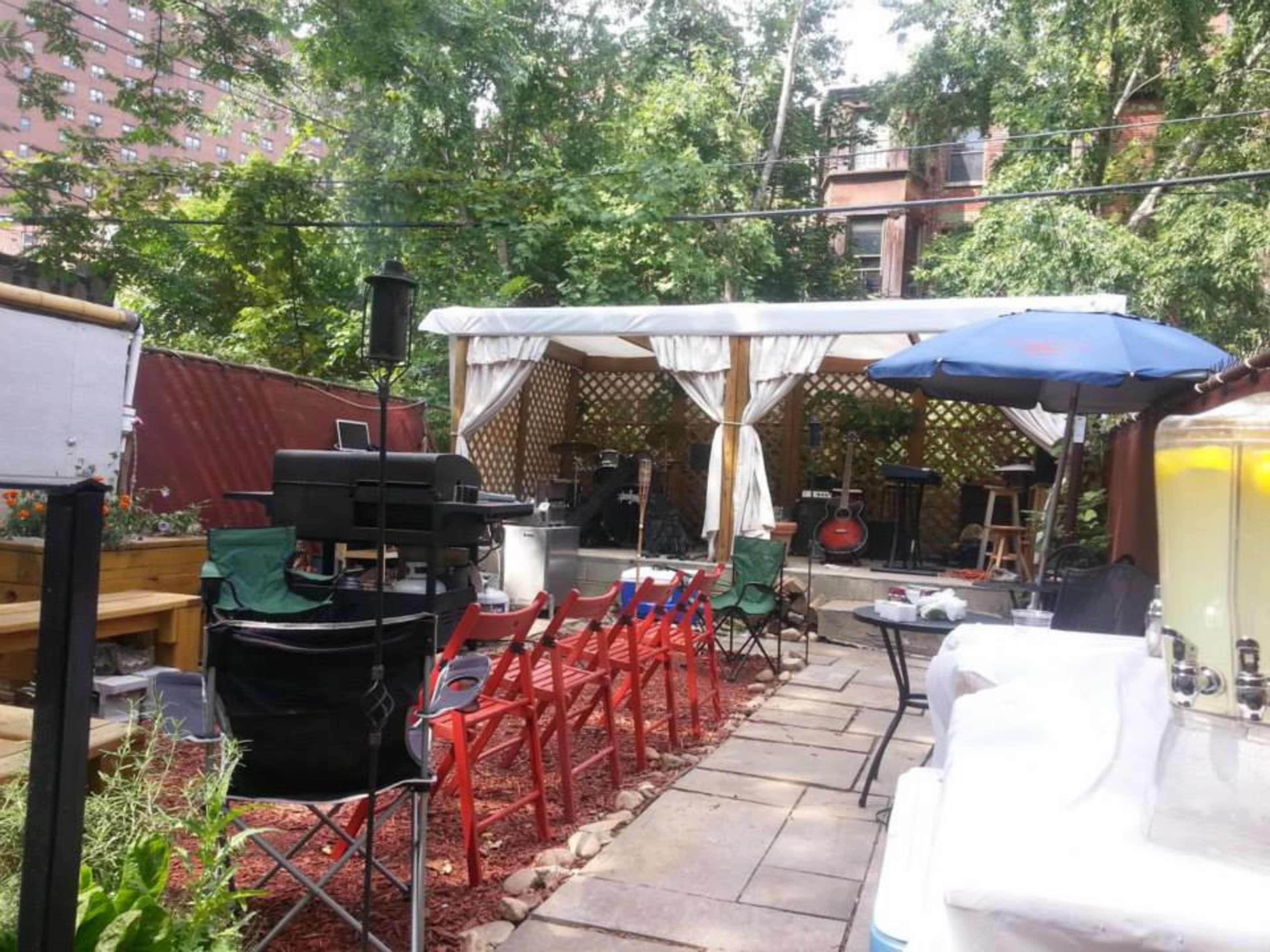 An outdoor event space with a covered area for music, surrounded by greenery and seating arranged with red chairs.
