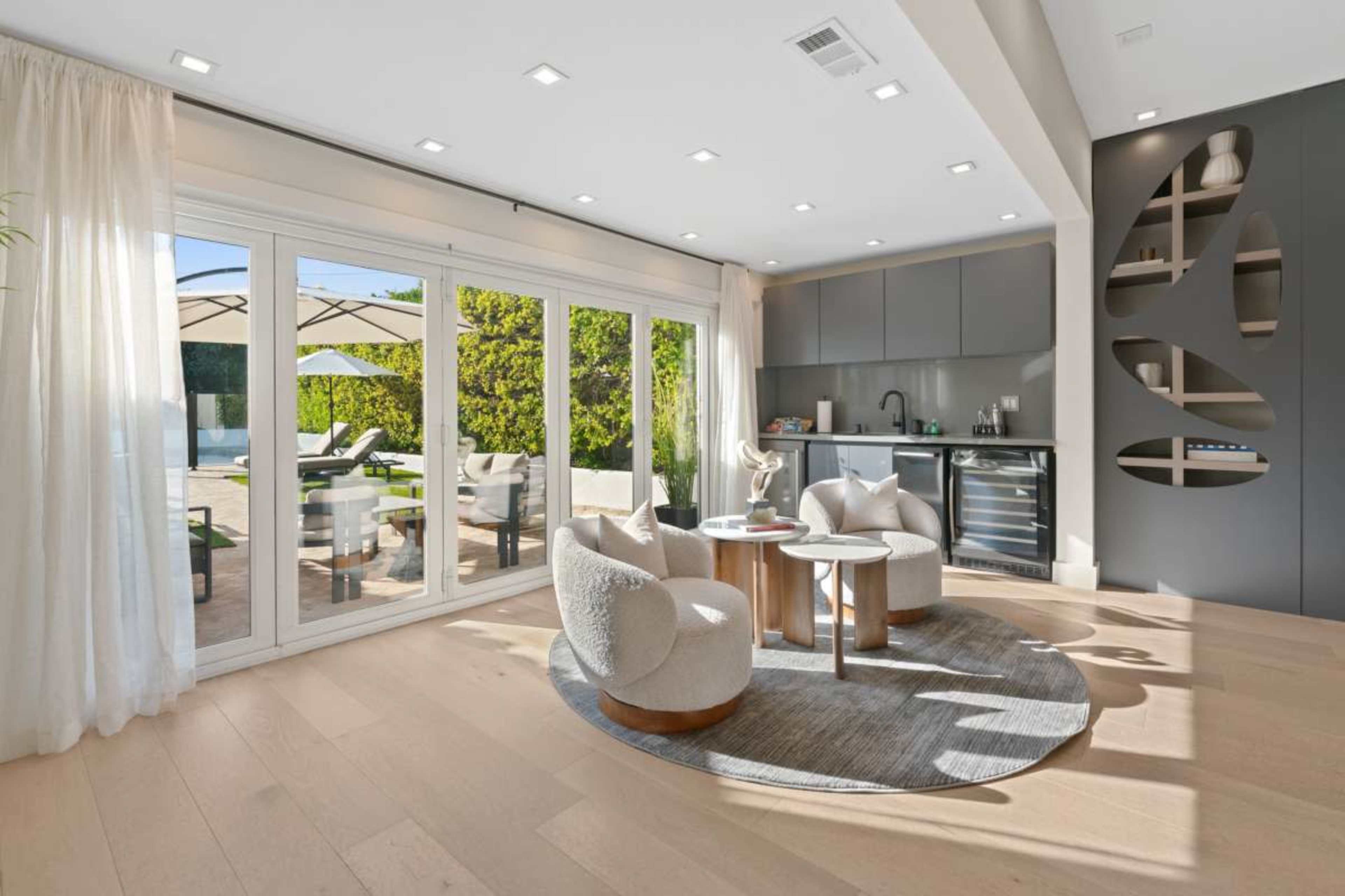 A bright living space features two circular armchairs and a round table on a rug, with large windows offering a view of a patio and outdoor seating area.