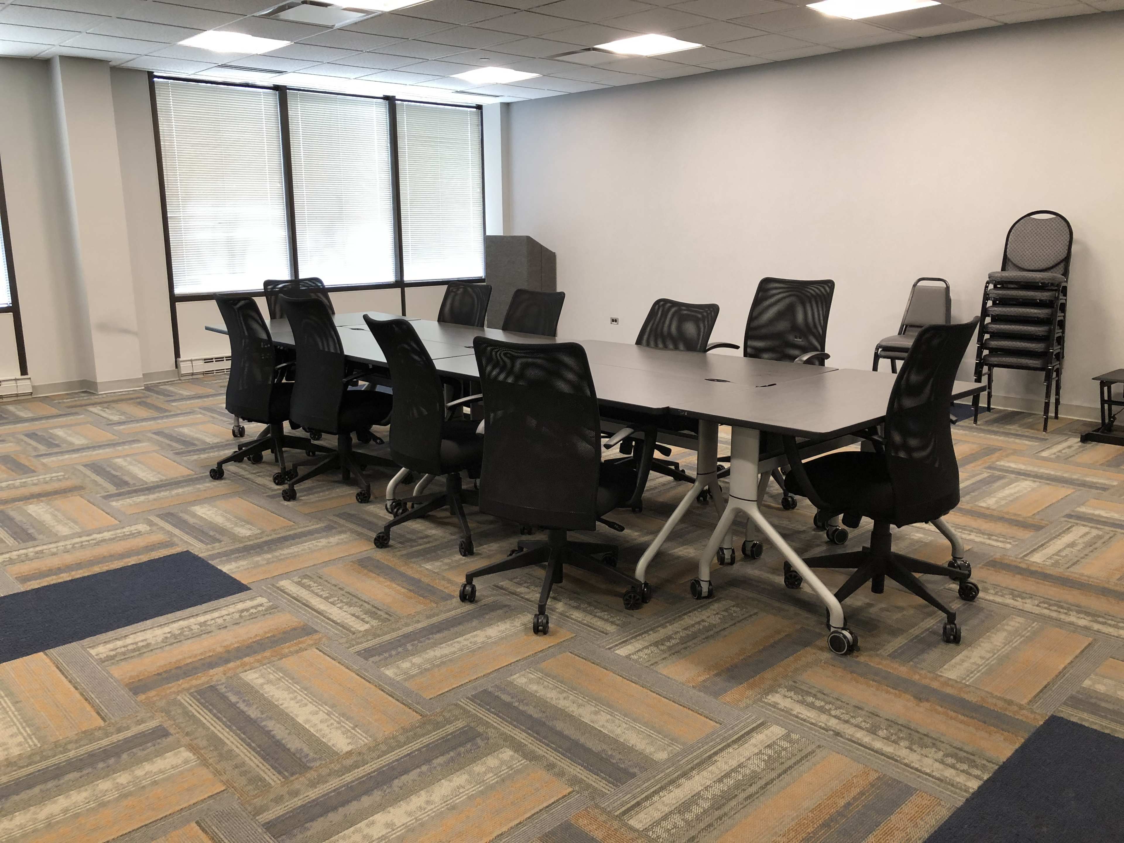 A conference room features a long table surrounded by black rolling chairs, with large windows on one side and a podium in the corner.