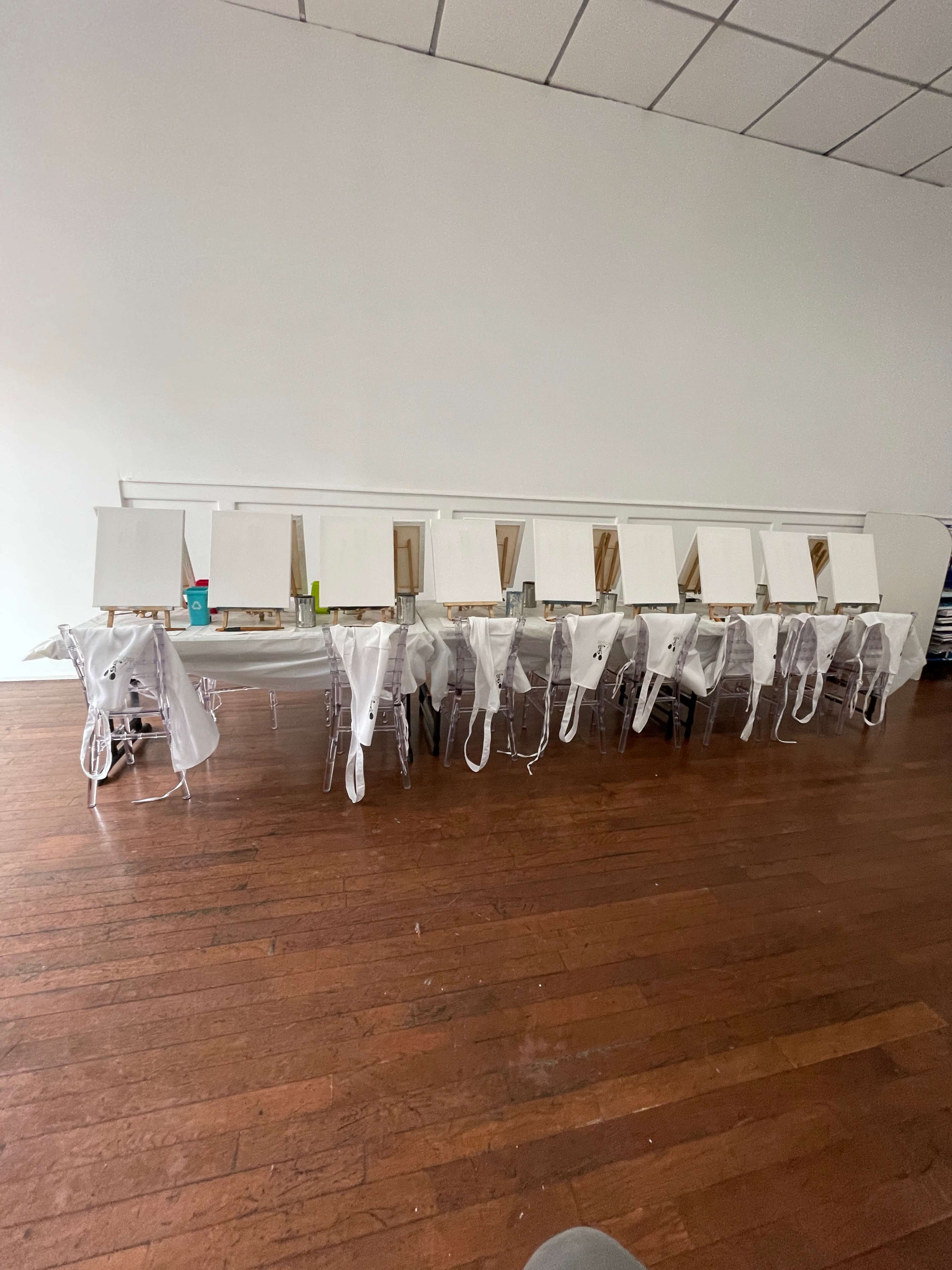 The image shows a row of empty easels with blank canvases set up on a long table in a spacious room with wooden flooring.