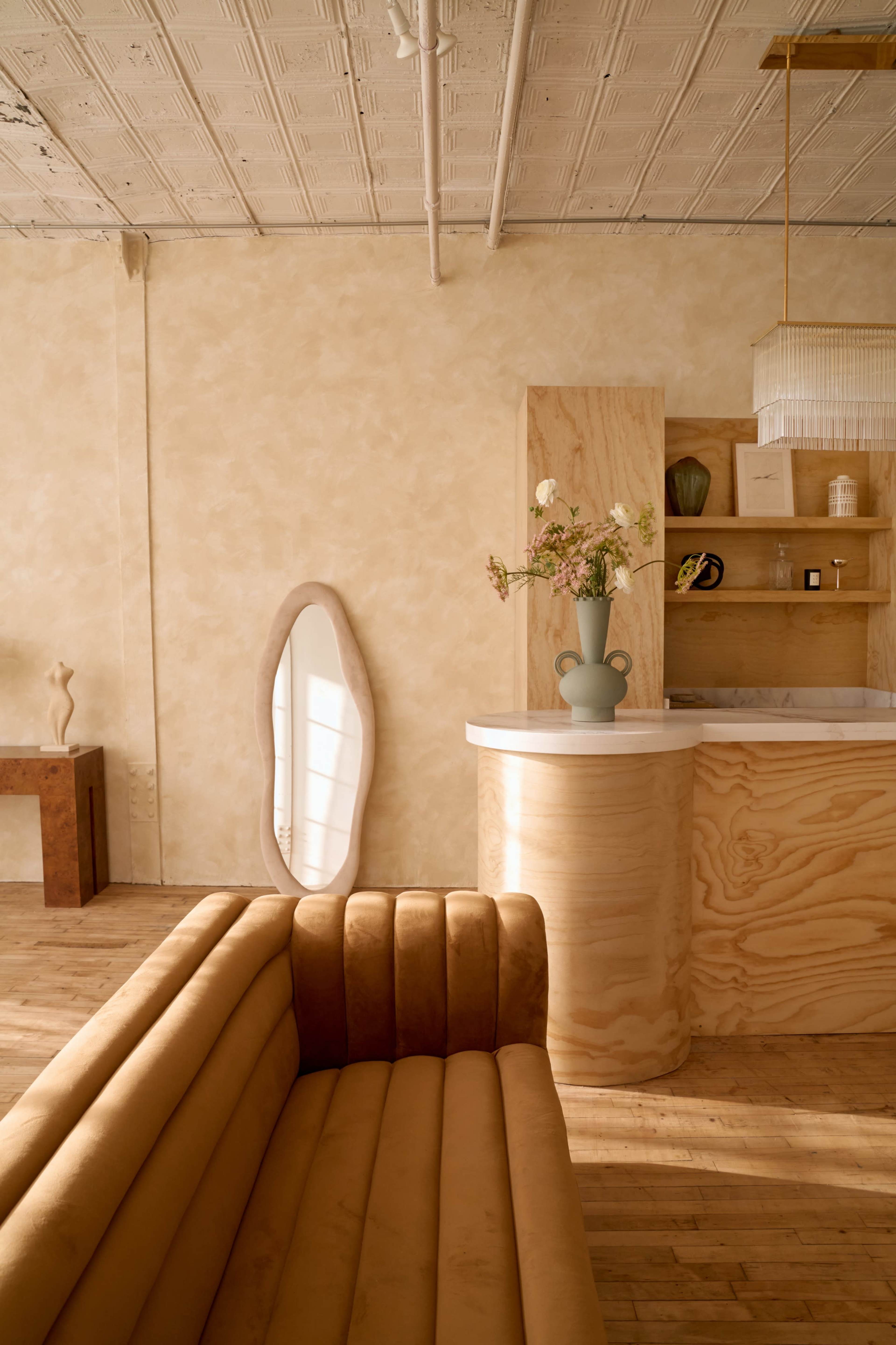 The image shows a modern interior with a curved tan sofa, a wooden counter, a decorative vase with flowers, and a large mirror against a textured wall.