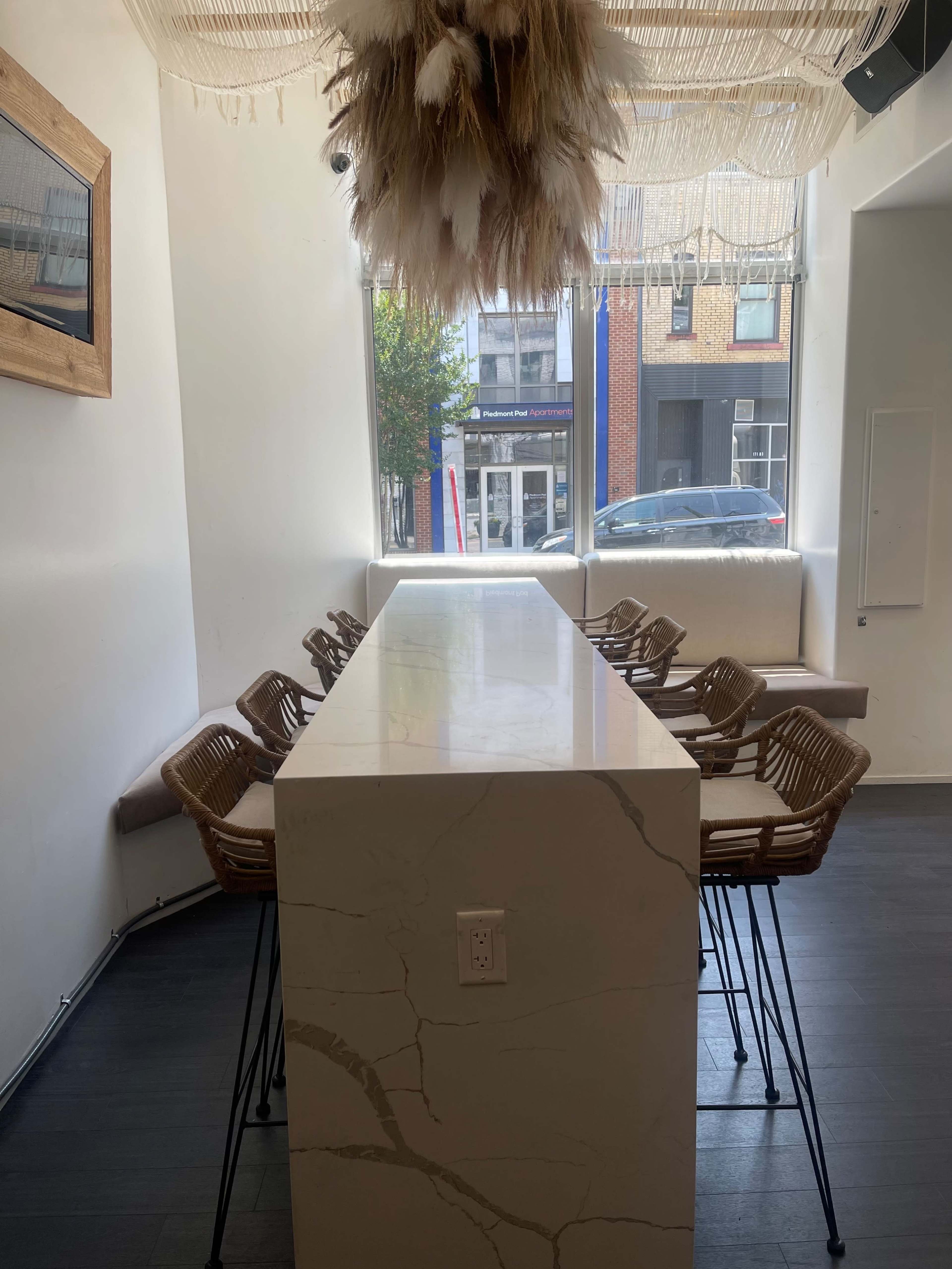 A long, marble table with wicker chairs is positioned in a white room featuring a large decorative light fixture overhead.