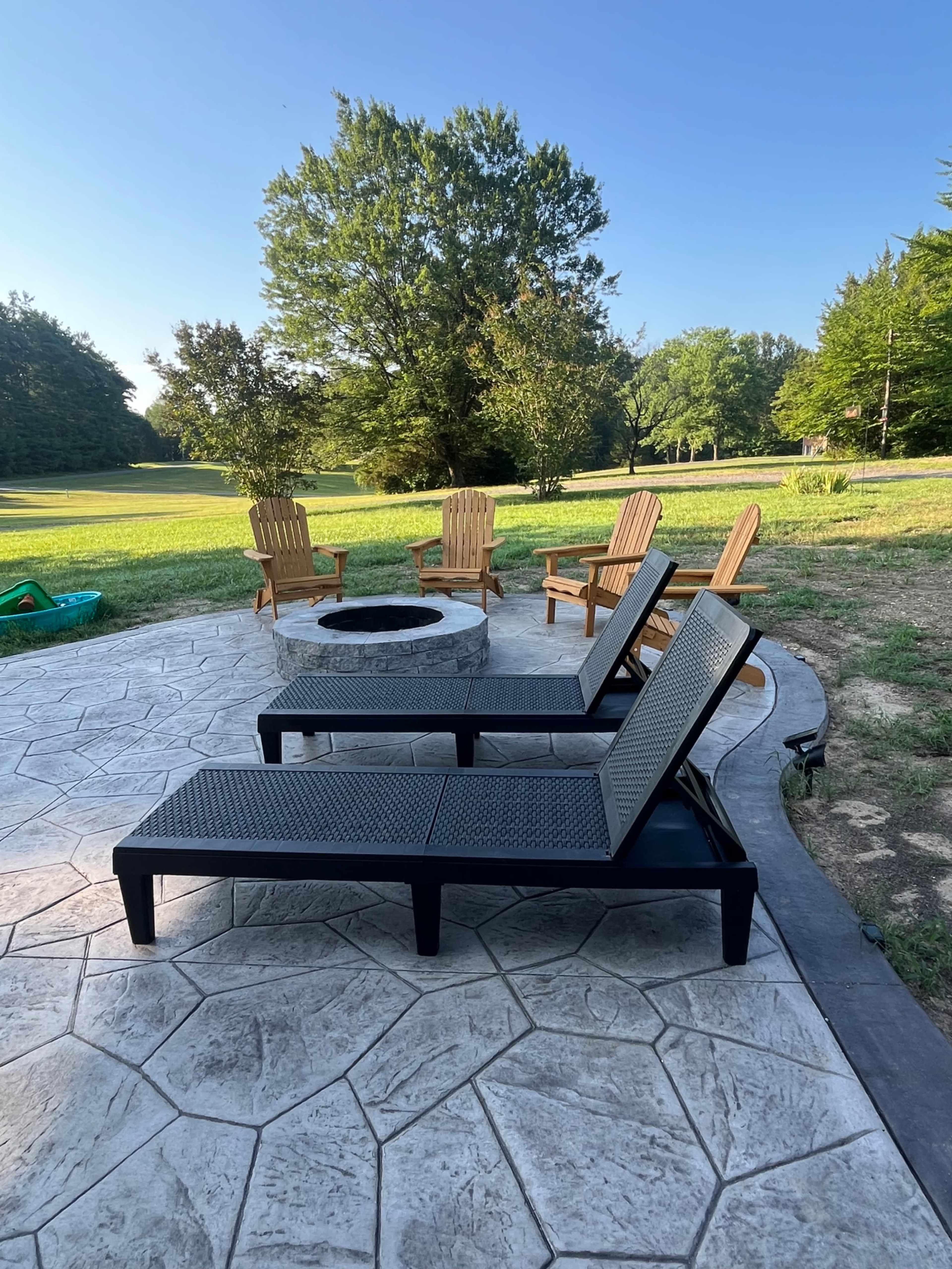 A circular fire pit is surrounded by four wooden chairs on a stone patio, with two lounge chairs positioned nearby in a grassy outdoor area.