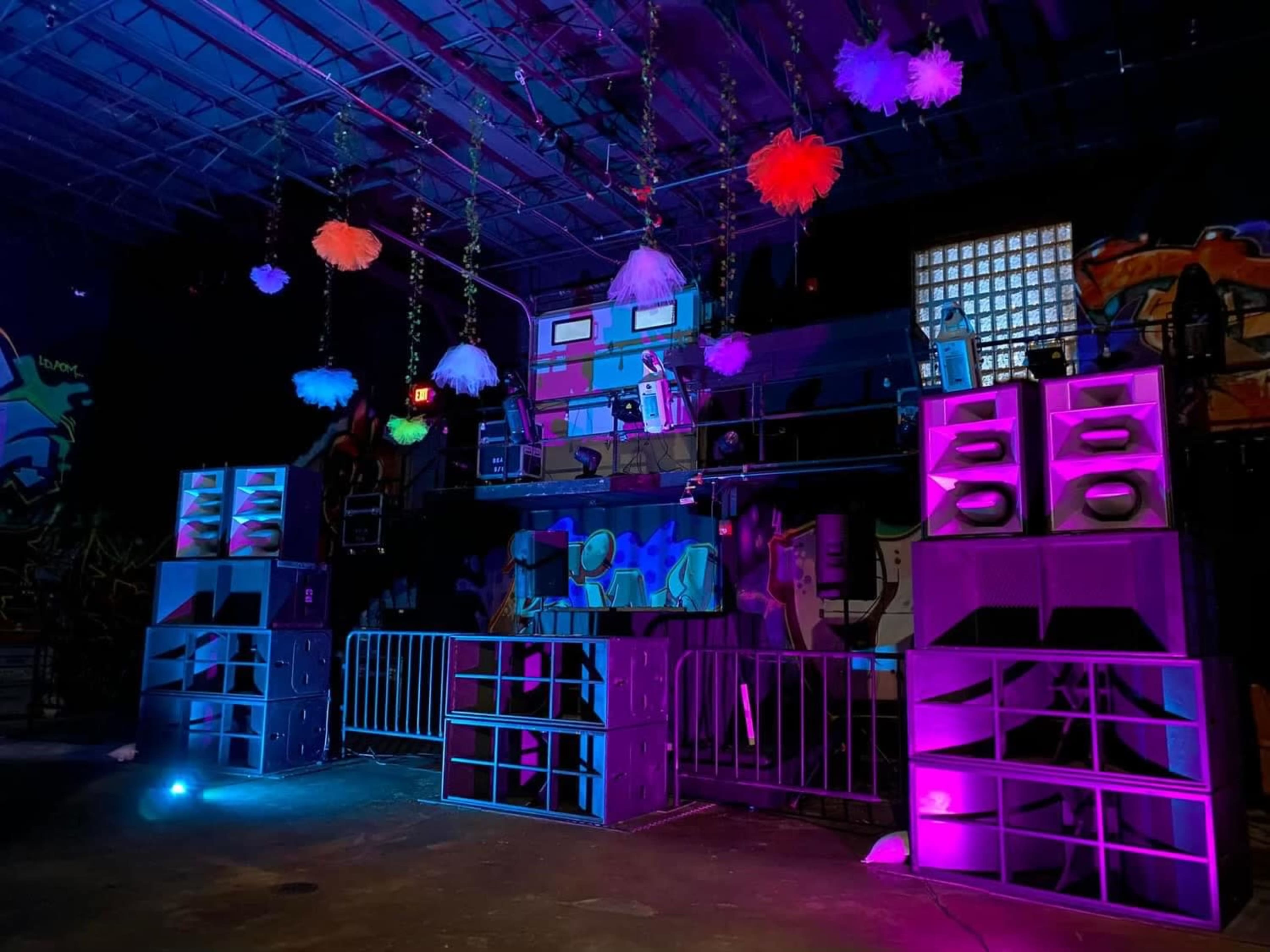 The image shows a dimly lit indoor venue set up for an event, featuring colorful speakers arranged in stacks and decorative hanging elements.