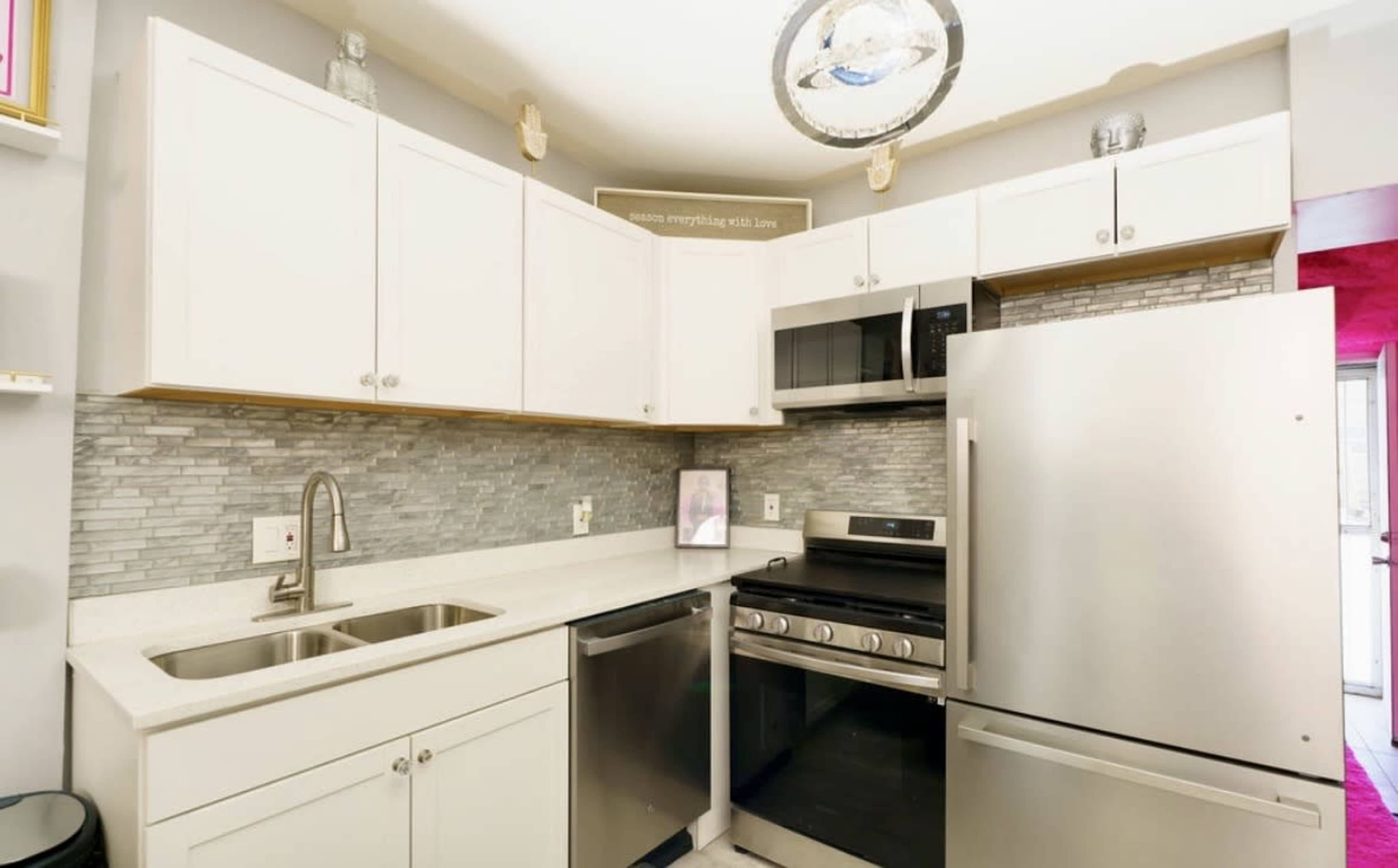 The image shows a compact kitchen featuring white cabinets, stainless steel appliances, and a mosaic tile backsplash.