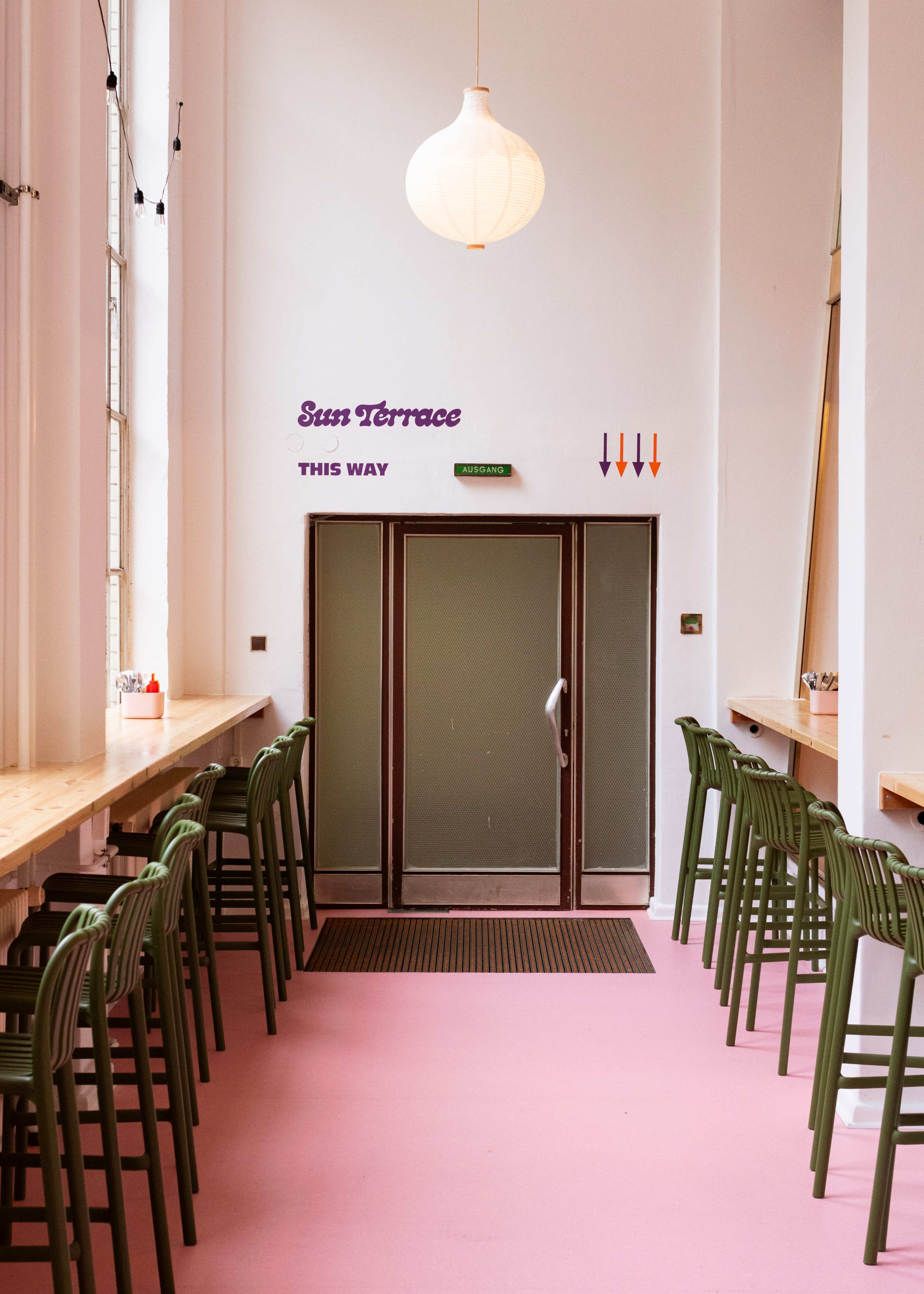 The image shows a brightly lit interior space with a series of green bar stools aligned along a wooden countertop, leading to a glass door labeled "Sun Terrace" with directional arrows above it.