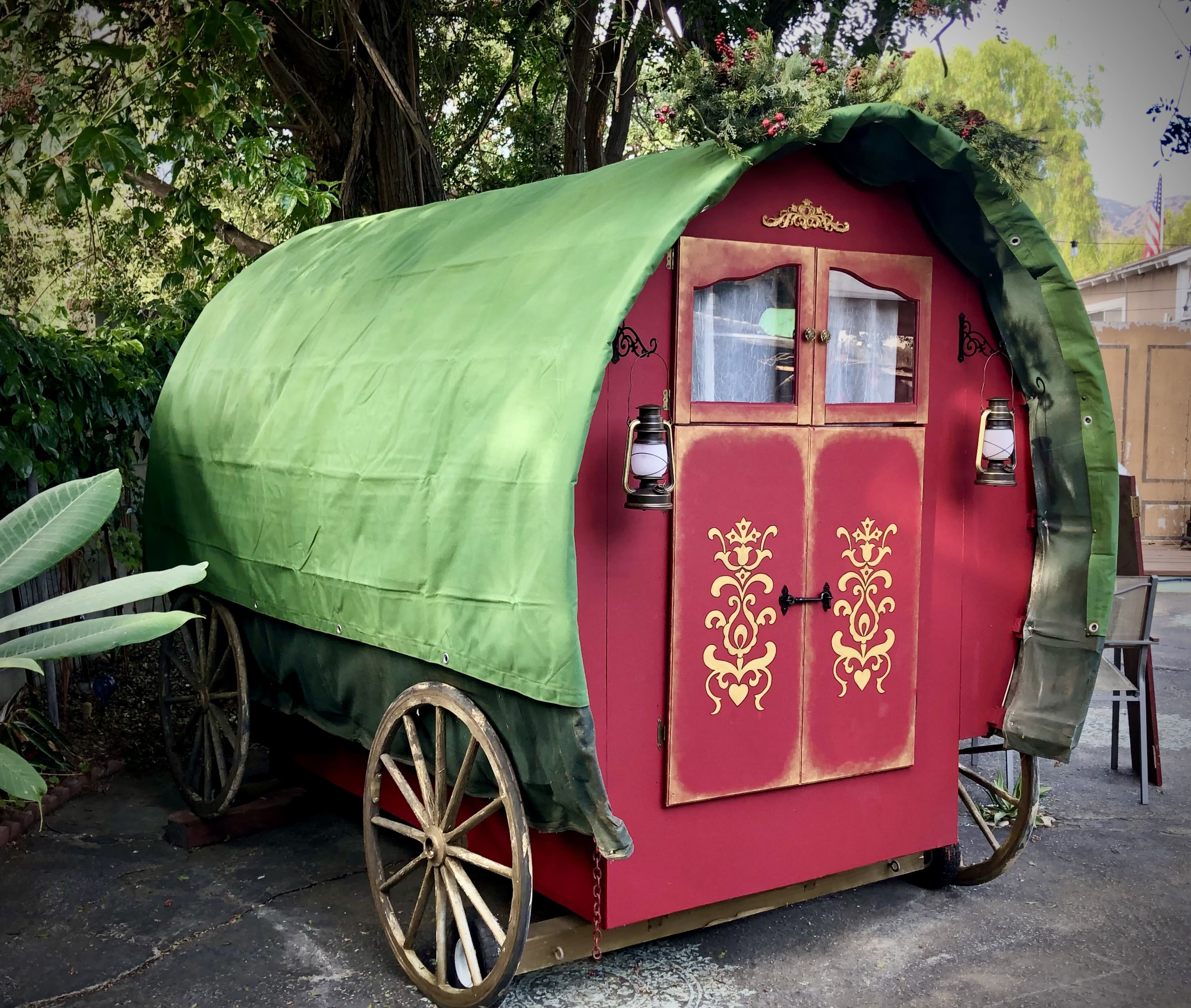 A colorful, mounted caravan with a green tarp roof and decorative door is parked in a yard surrounded by trees.