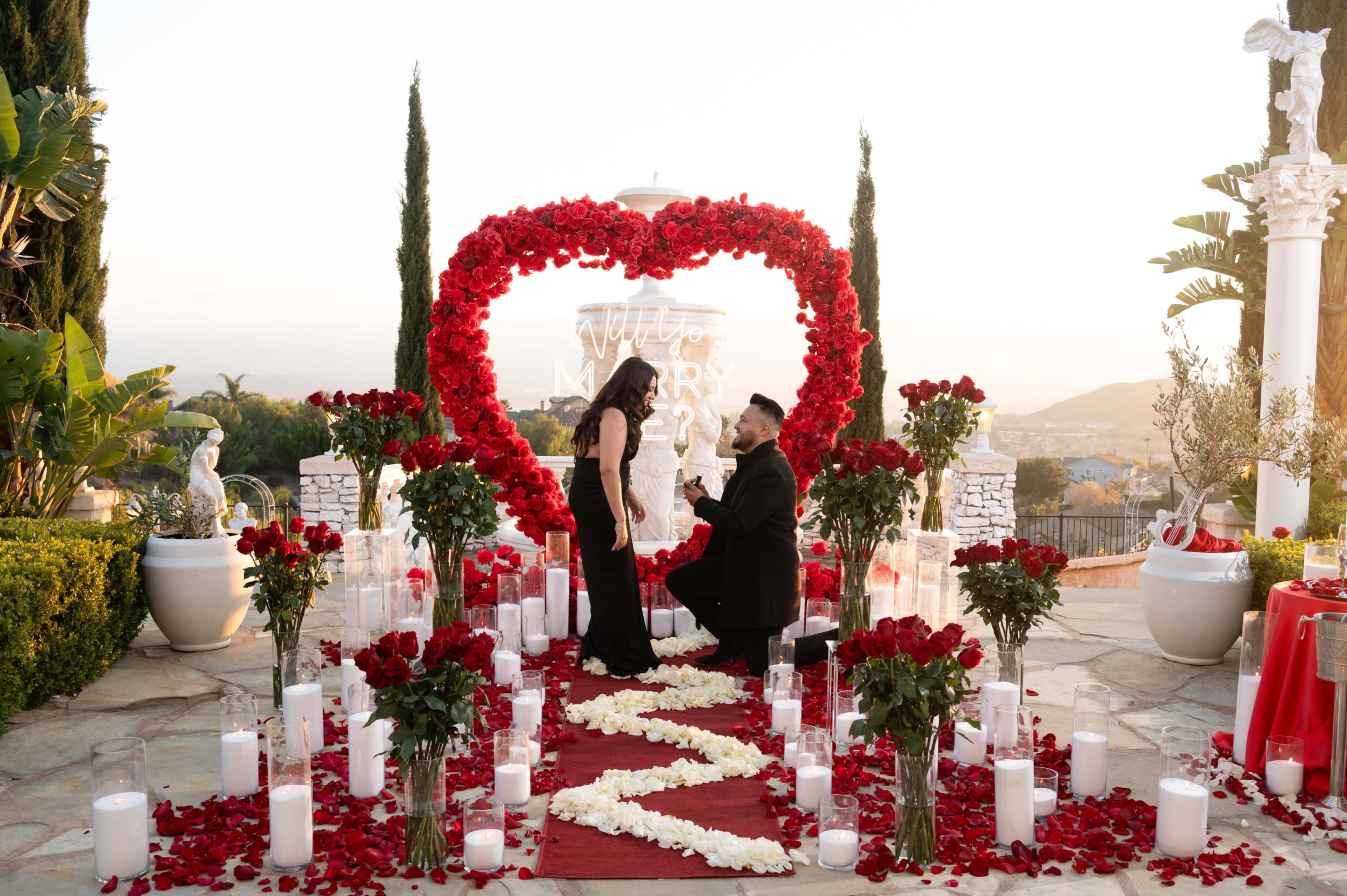 A man is kneeling in front of a woman in a floral decorated area, surrounded by rose petals and candles, with a large heart made of roses in the background.