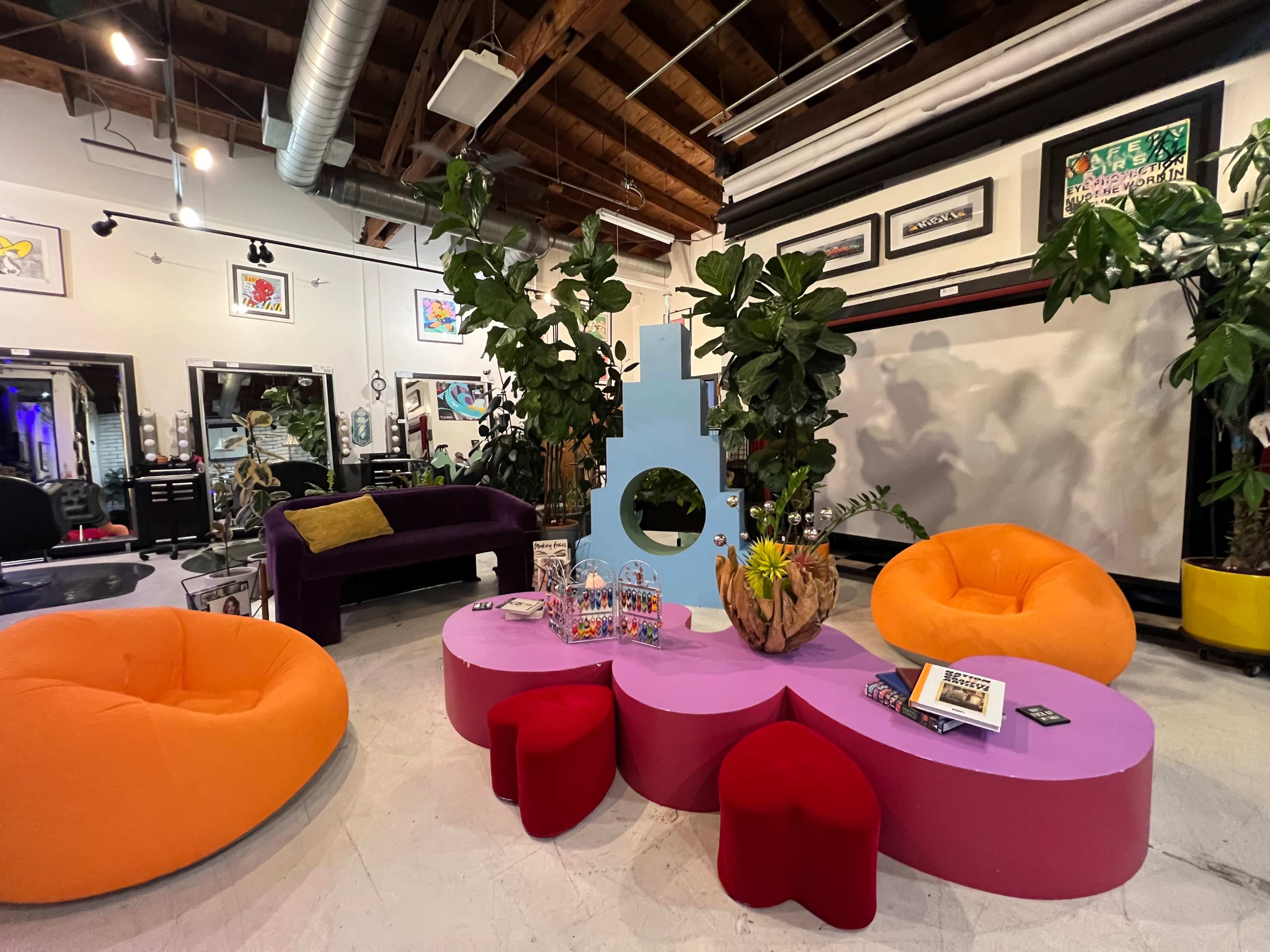 The image shows a modern, stylish lounge area featuring colorful furniture including orange and pink beanbag chairs, a purple sofa, and a flower-shaped coffee table surrounded by indoor plants.
