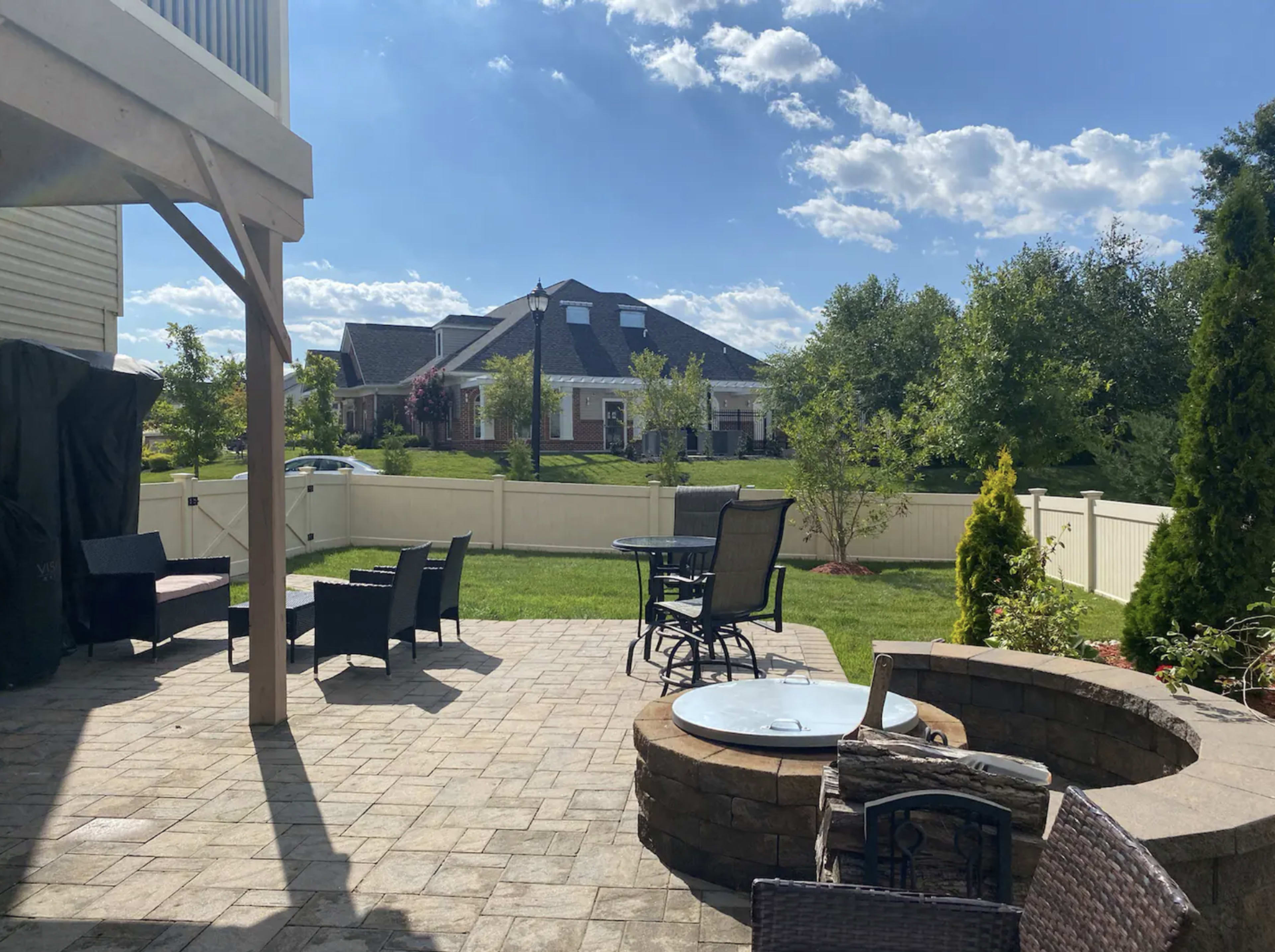 The image shows a patio area with seating and a small fire pit, surrounded by a well-maintained lawn and a house in the background under a partly cloudy sky.