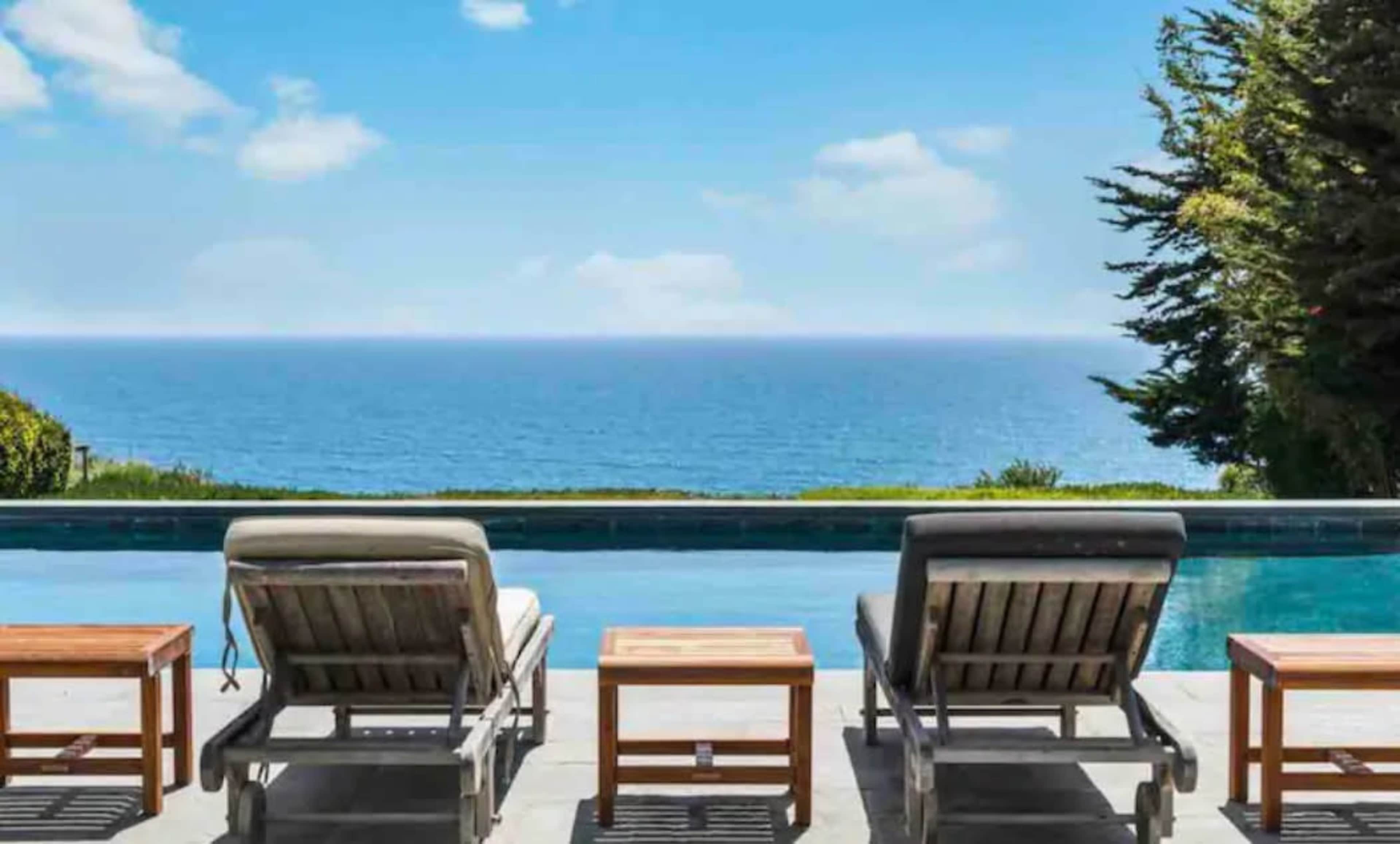 A pool area with two lounge chairs facing the ocean, with a clear sky and lush greenery in the background.