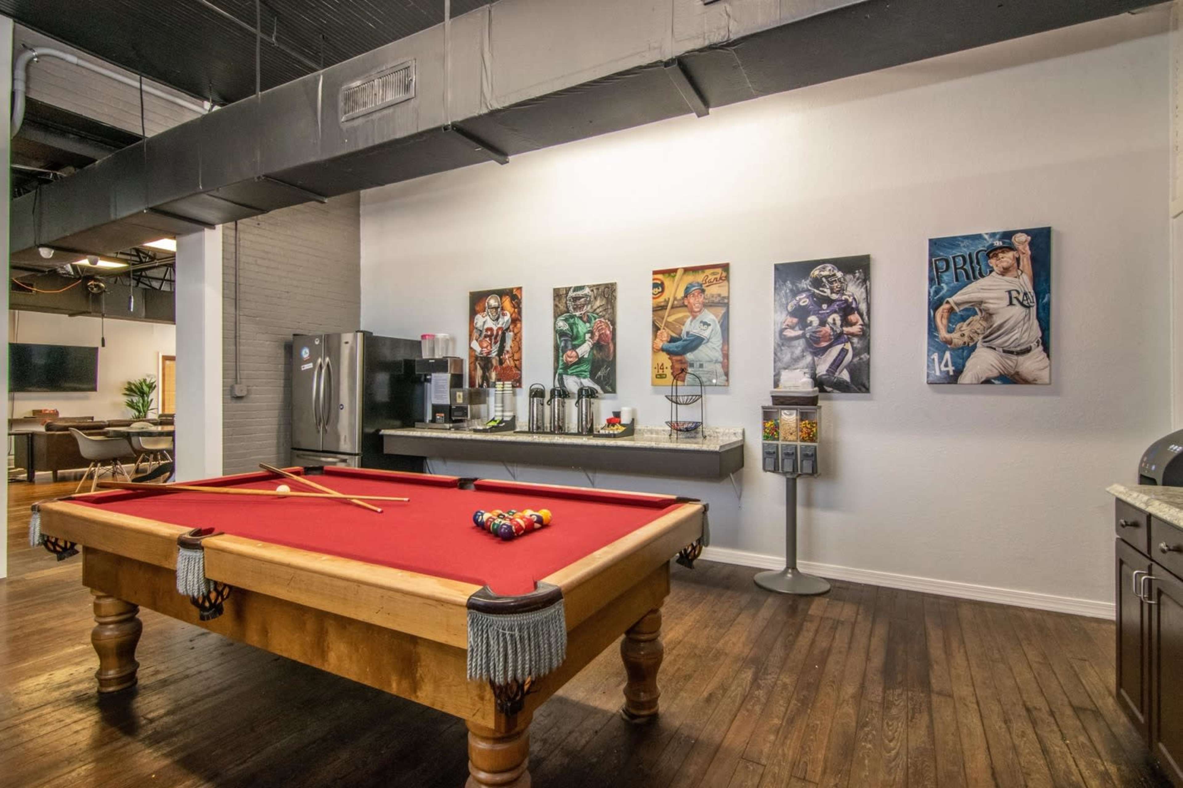 A recreational room featuring a red billiard table with cues, surrounded by sports-themed artwork on the walls and a refreshment station in the background.
