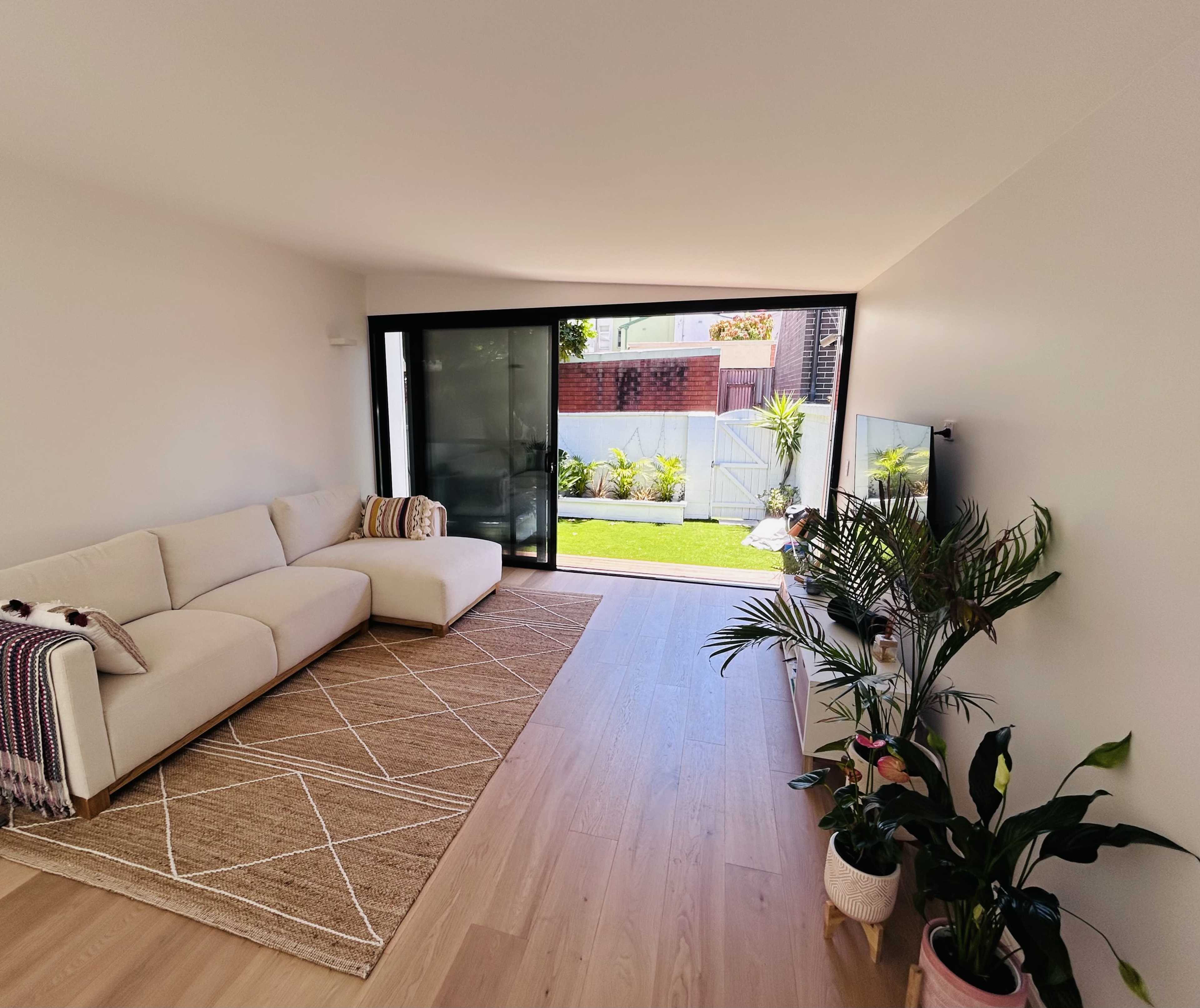 The image shows a bright living room with a white couch, a large window opening to a garden, and indoor plants.