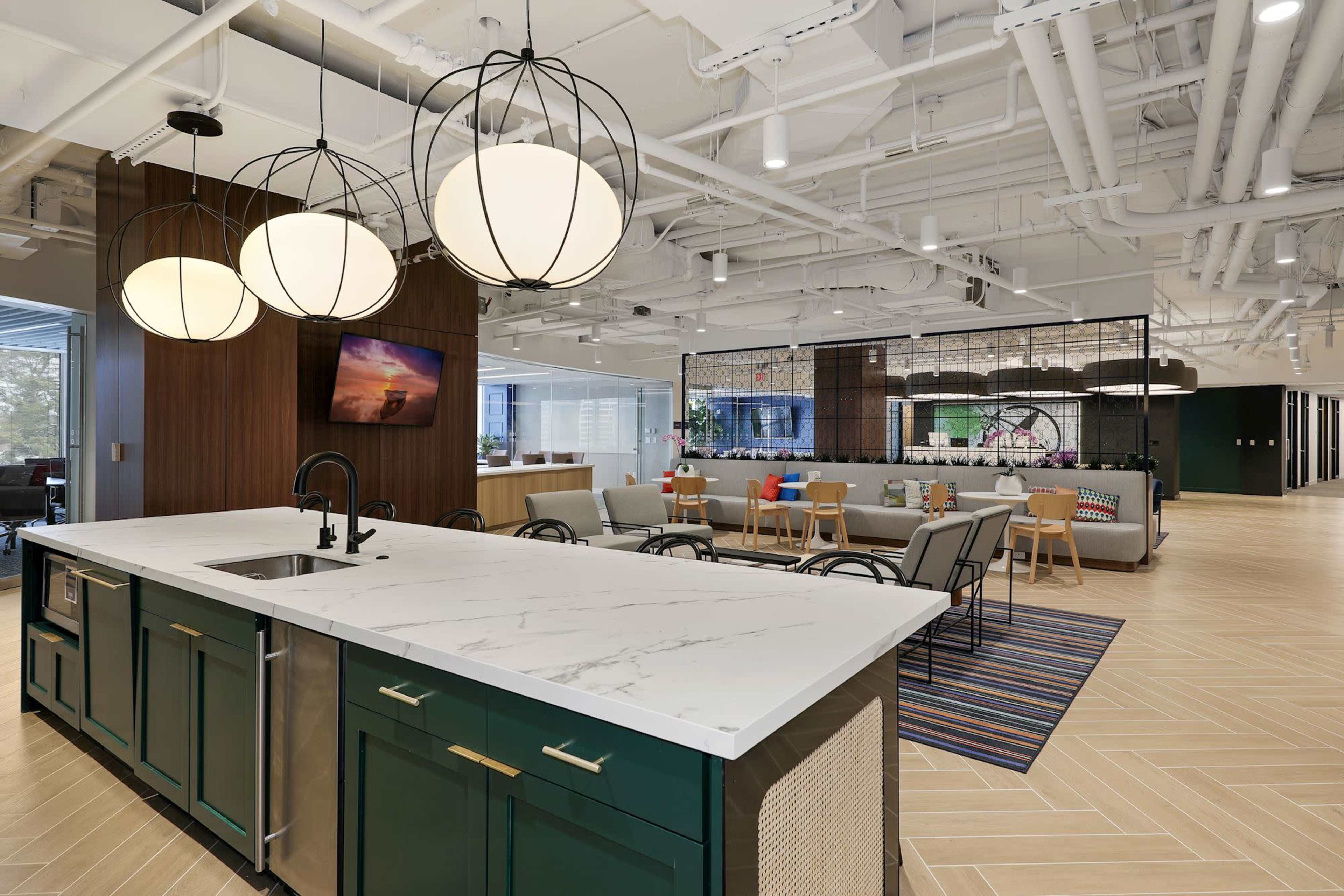 The image shows a modern office kitchen with a large island, pendant lighting, a seating area, and a combination of wood and metal furnishings.