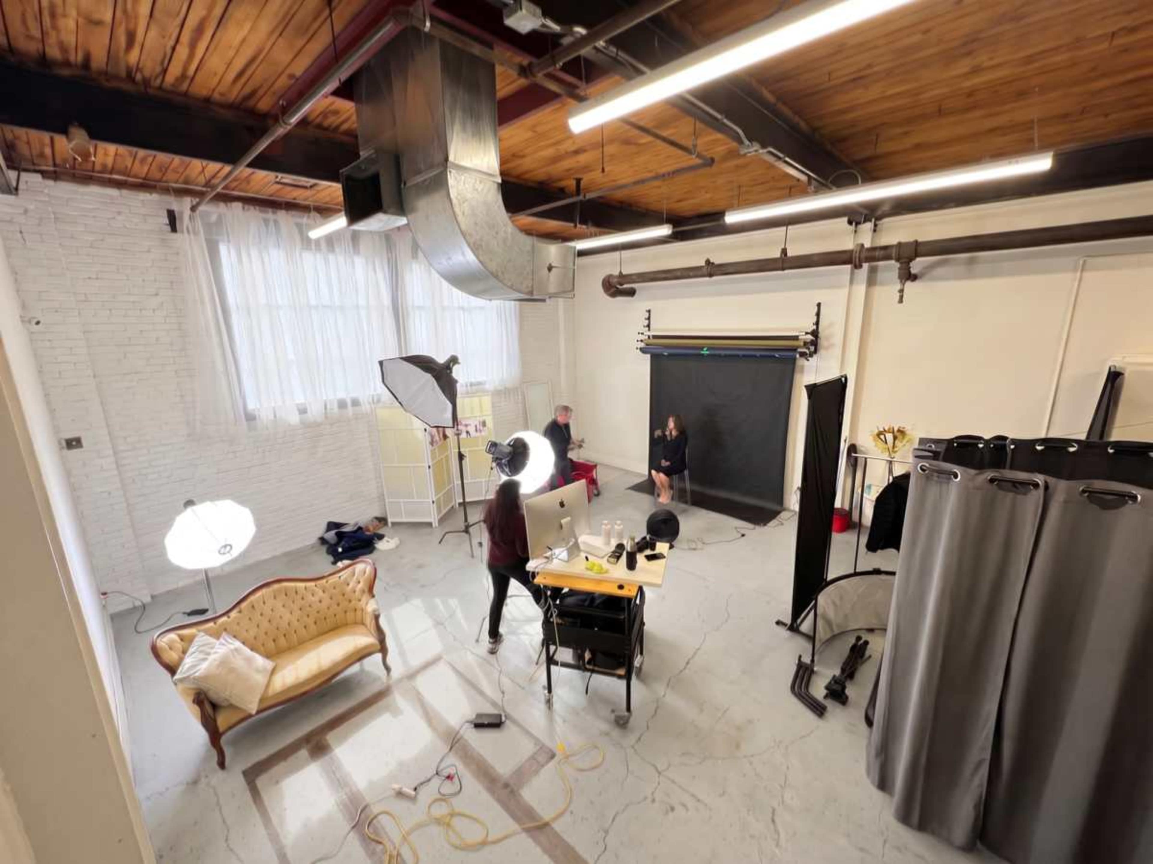 The image shows a spacious photography studio with a large window, two softbox lights, a backdrop setup, a table with equipment, and a vintage sofa.