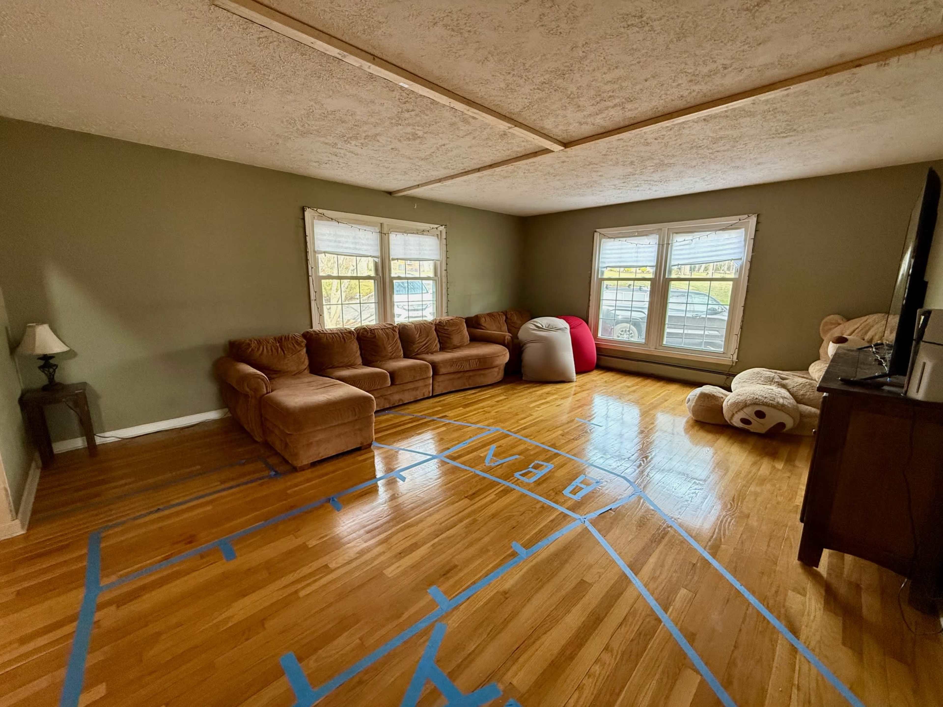 The room features a brown sectional sofa and a large teddy bear, with basketball court markings on the hardwood floor.