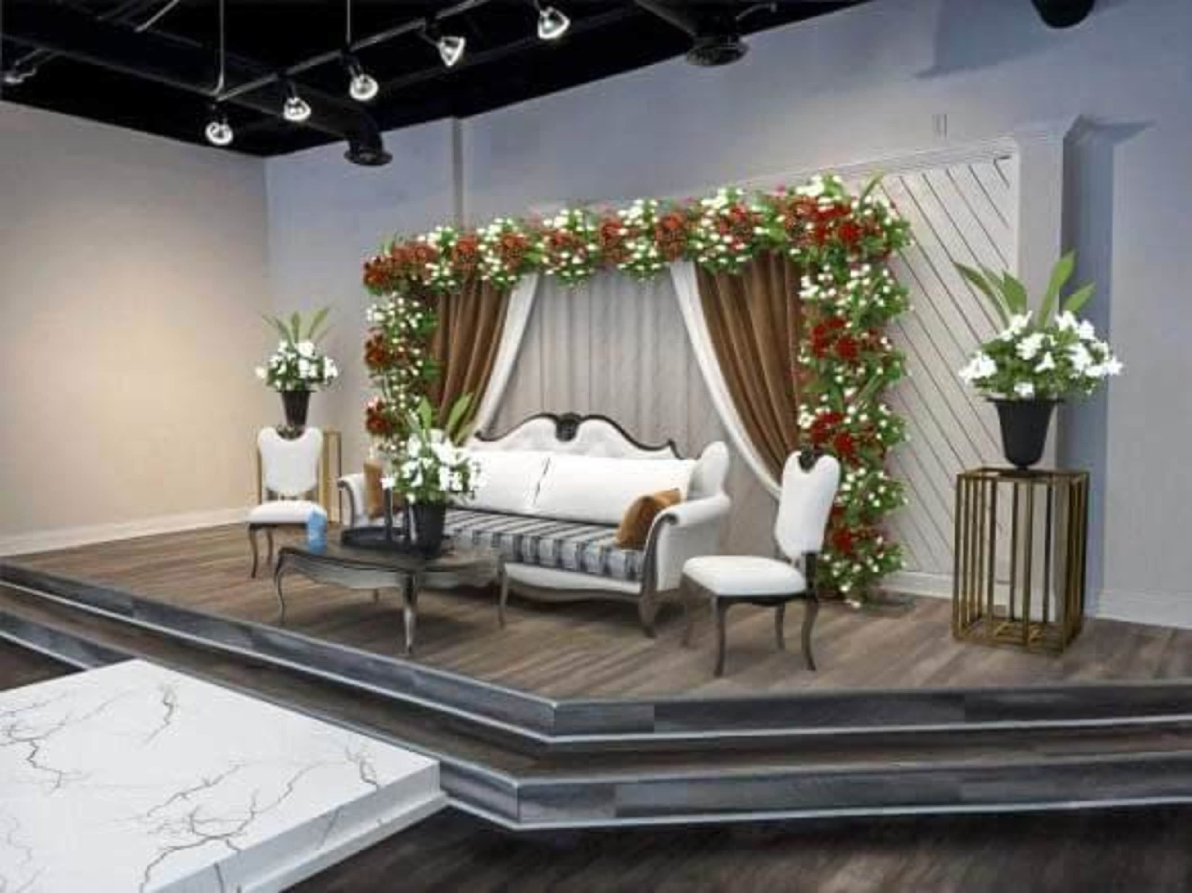 A decorated stage with a floral arch, a sofa, two chairs, and potted plants, set in a modern interior space.