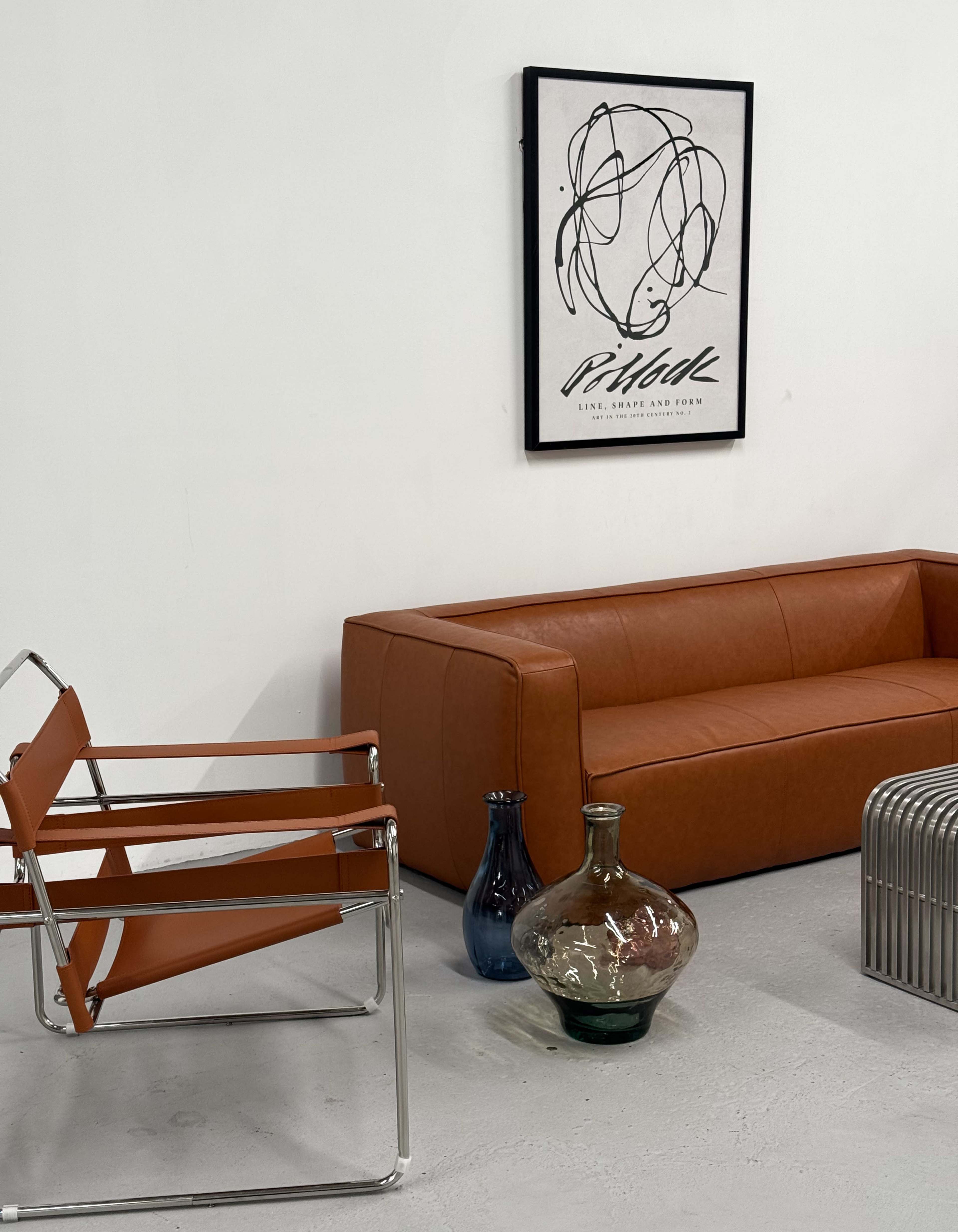 The image shows a modern interior featuring a brown leather sofa, a metal chair, and various decorative vases against a white wall with abstract art.