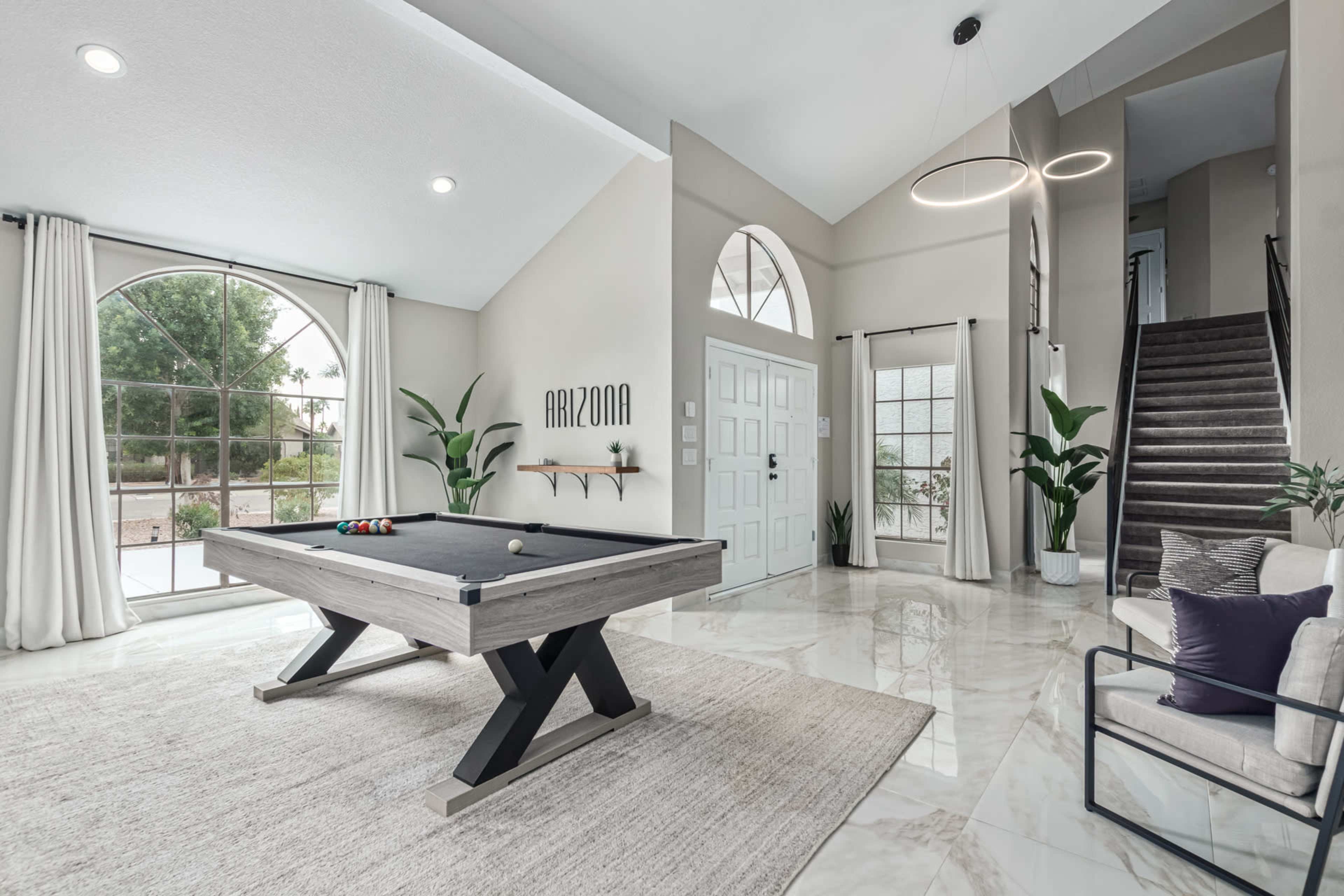 A spacious living area with a pool table, high ceilings, and large windows, complemented by potted plants and a staircase.