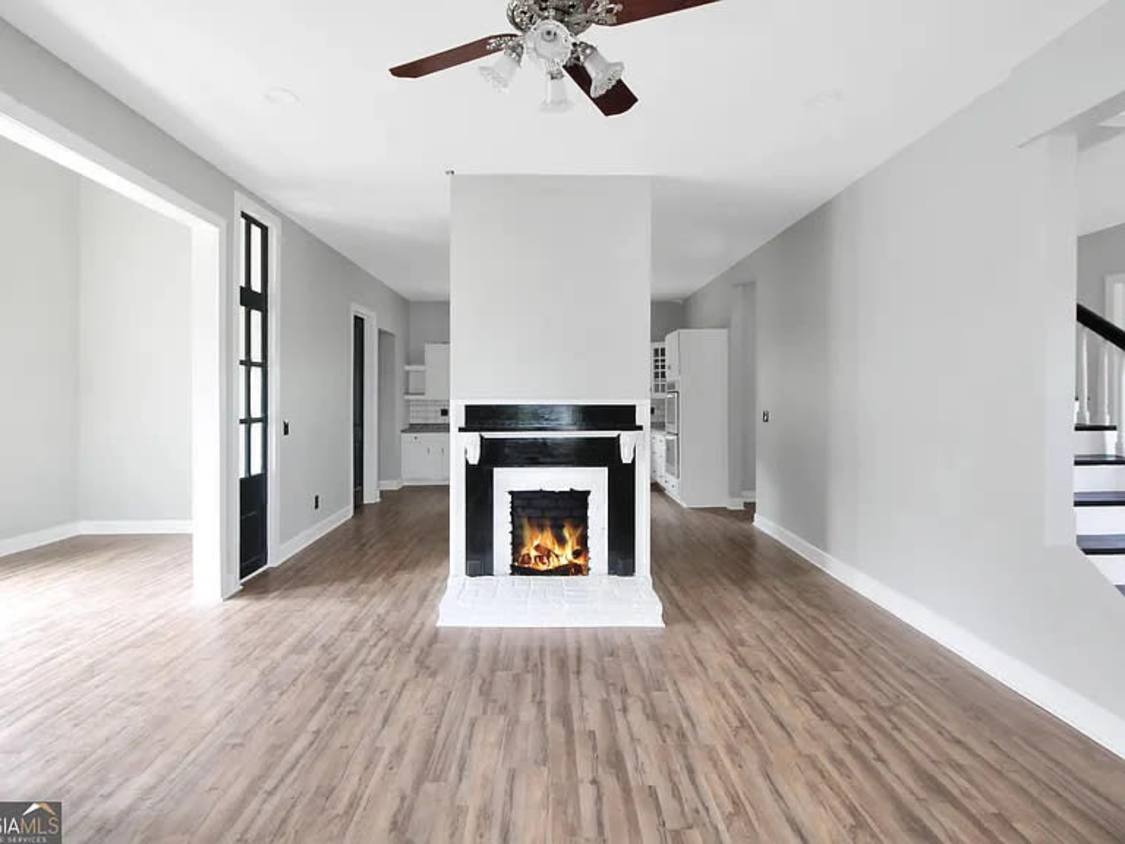 The image shows a spacious living room with a central white fireplace, light wood flooring, and gray walls.