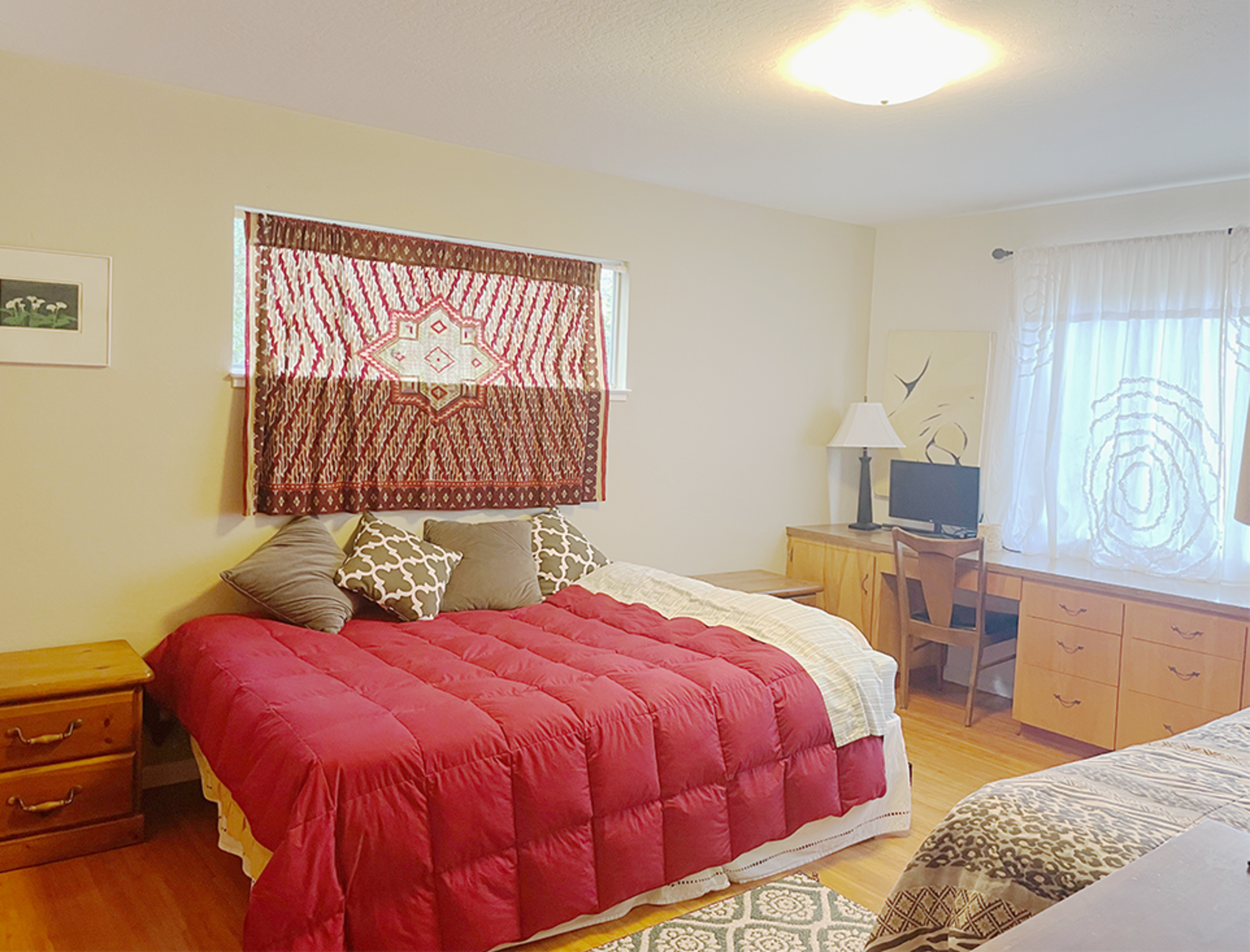 A bedroom with a large red bedspread, wooden furniture, a lamp, and a window covered with a sheer curtain.