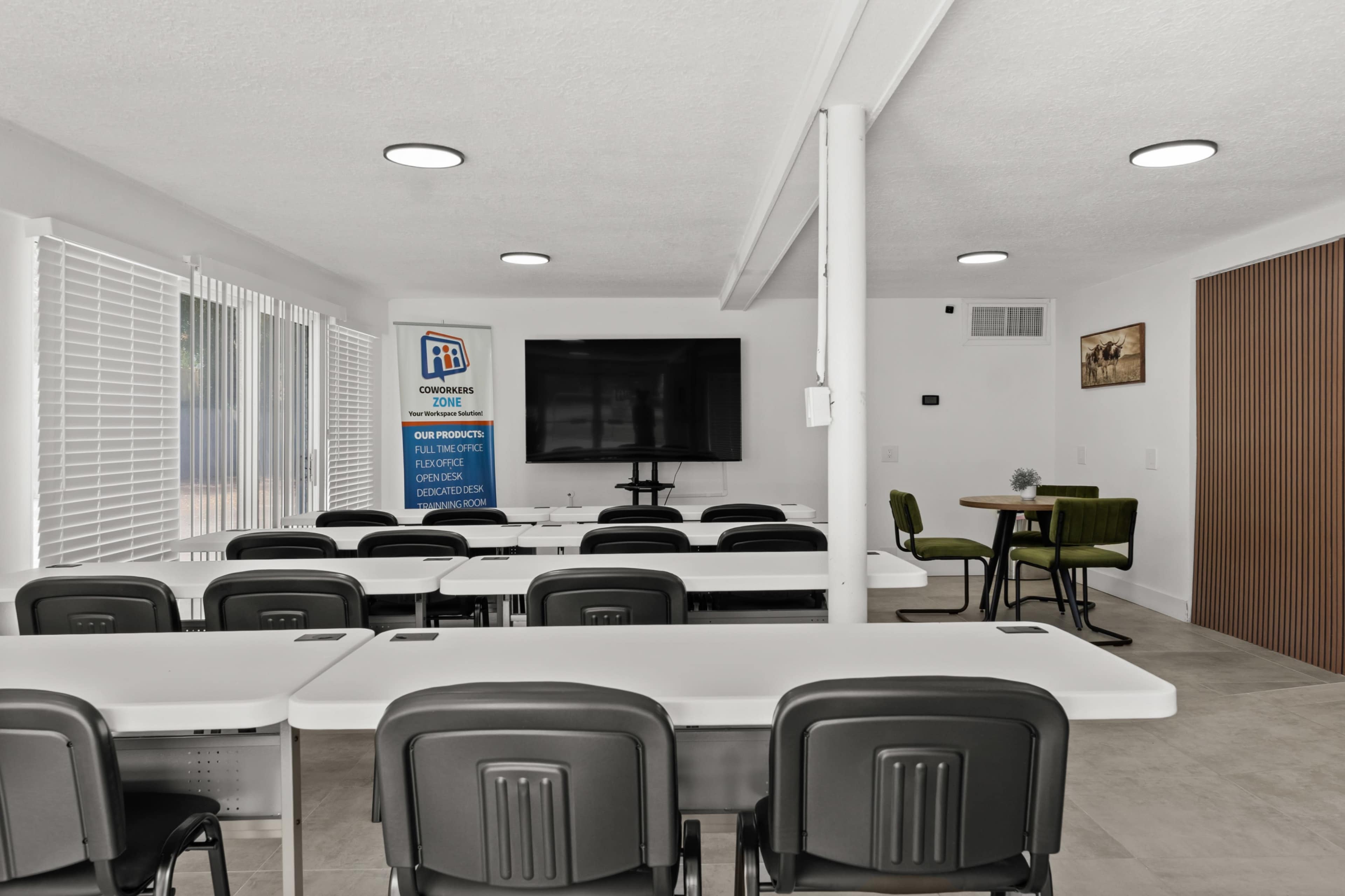 The image shows a modern conference room with rows of white tables and black chairs, featuring a large screen at the front and a small seating area in the corner.