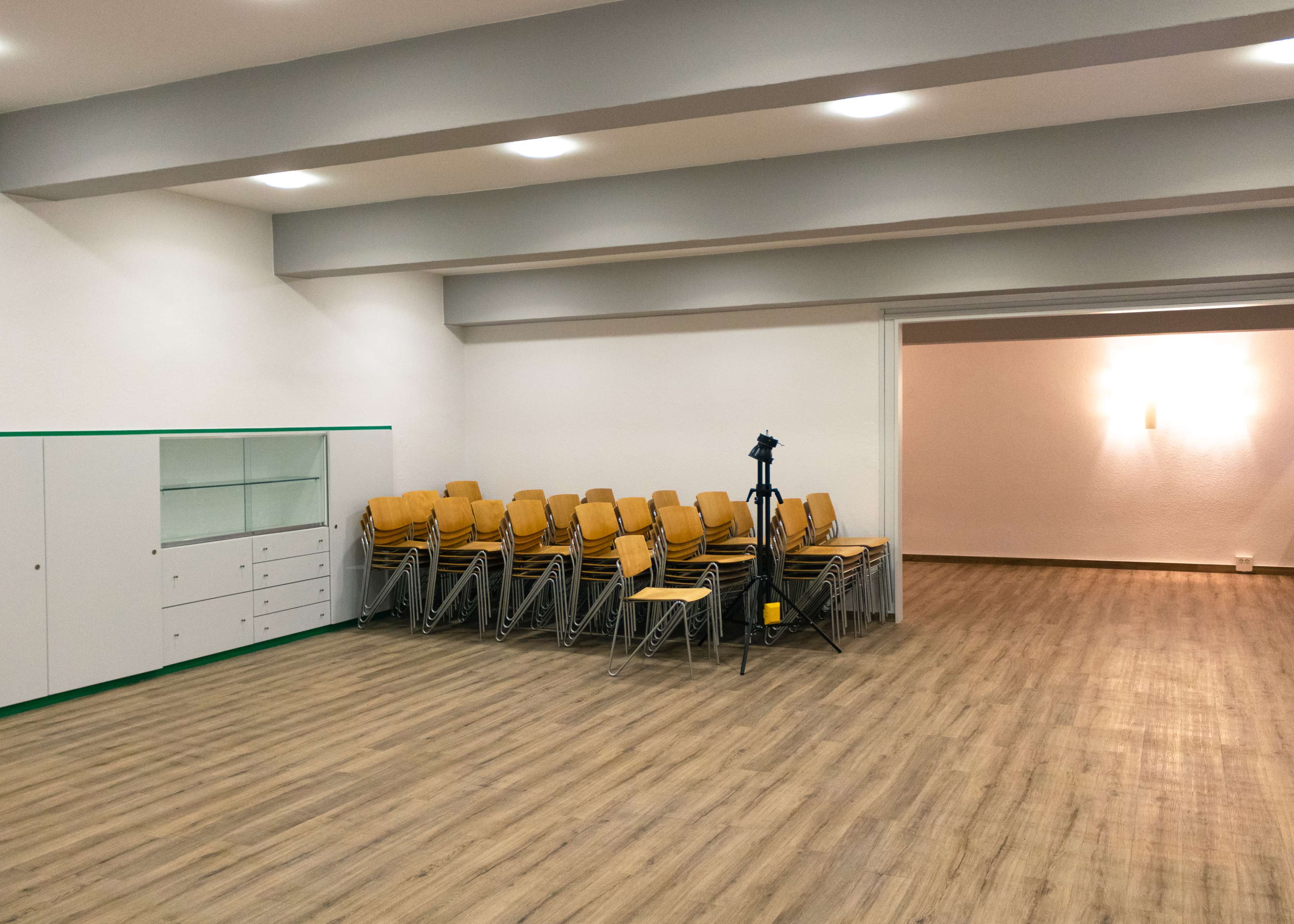 The image shows a spacious room with wooden flooring, featuring a stack of metal-framed chairs and a camera tripod positioned in one corner.