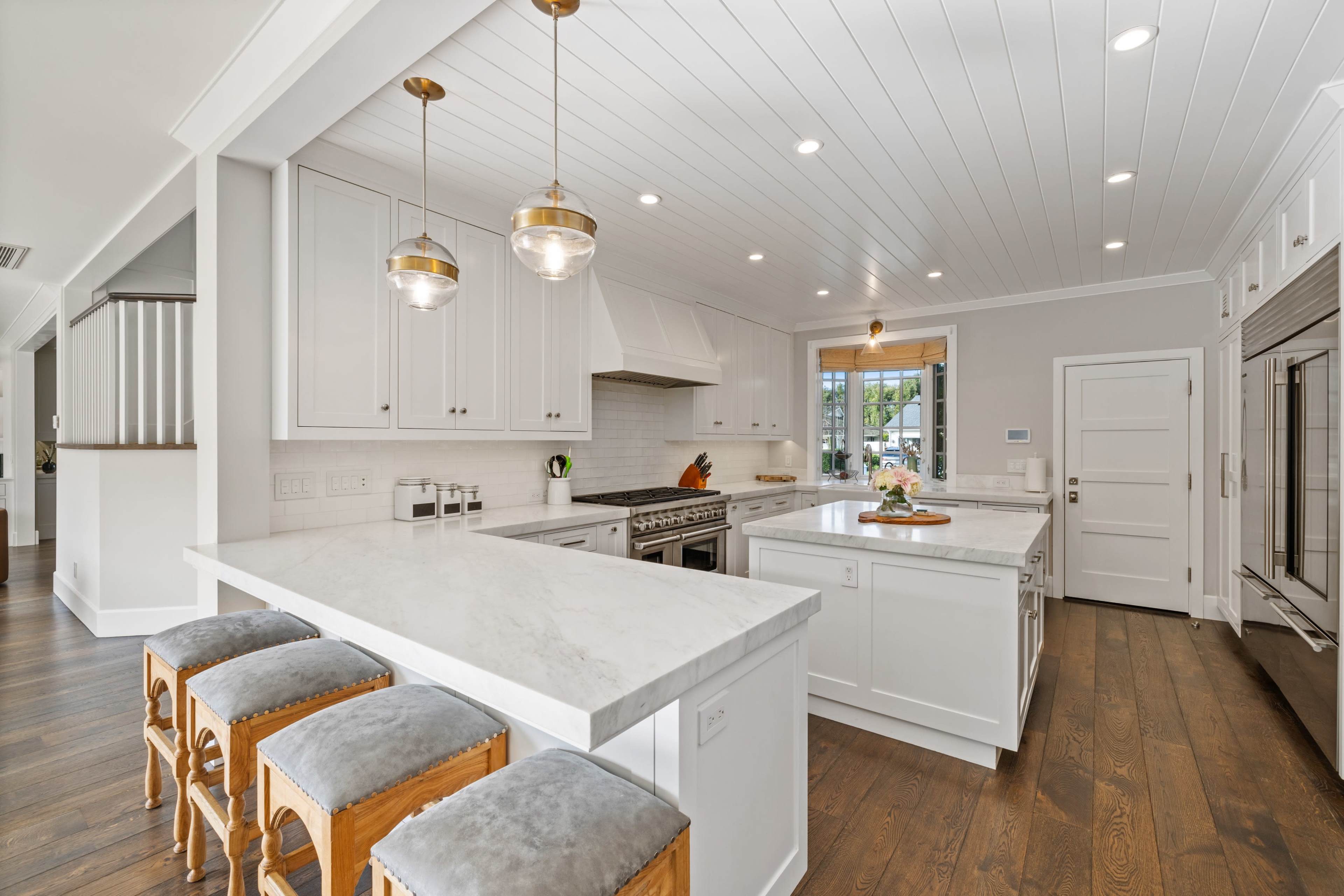 A modern kitchen features white cabinetry, a large marble island, and stainless steel appliances, with pendant lights overhead and a window letting in natural light.