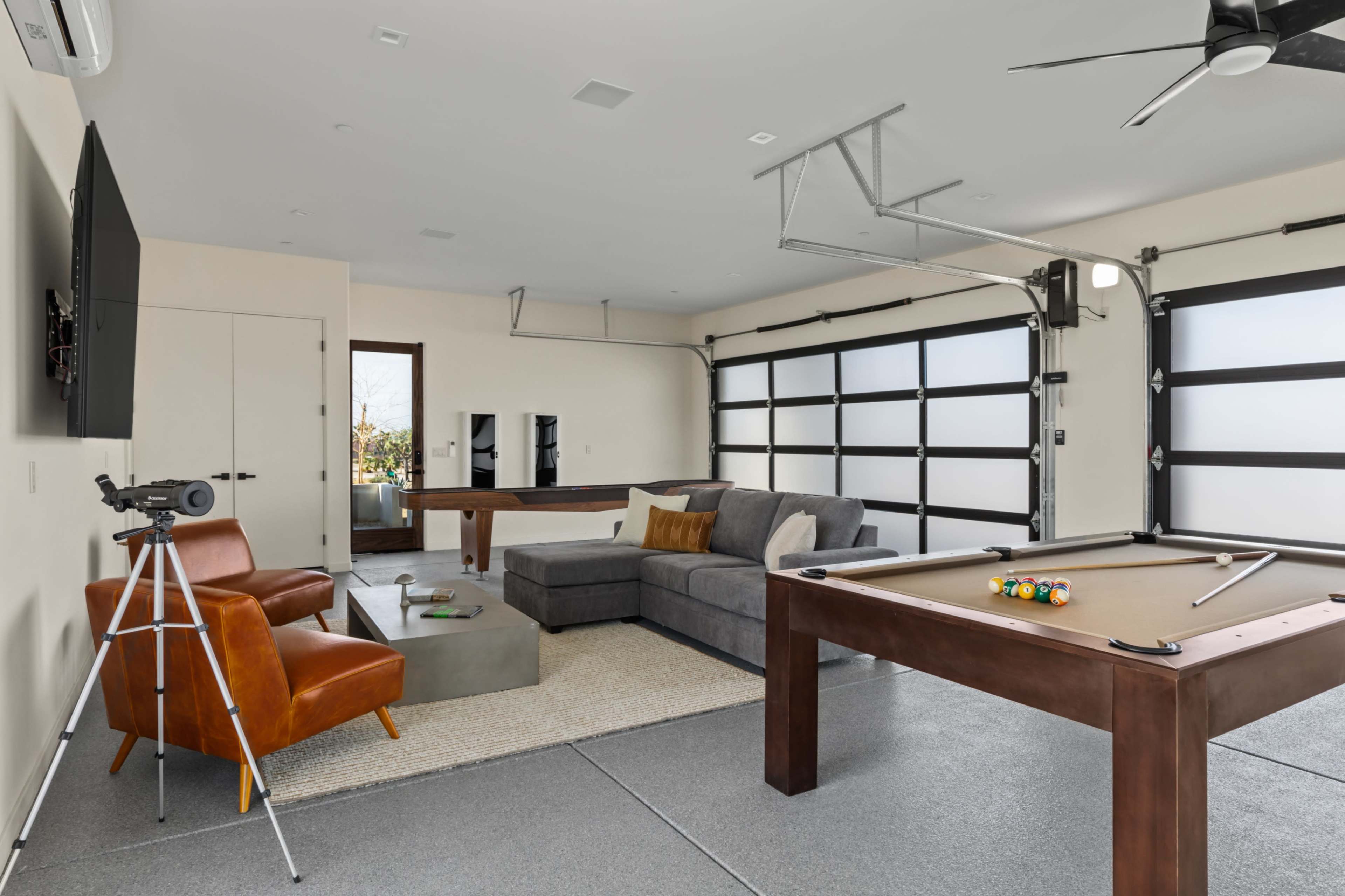 The image shows a modern garage converted into a recreational space with a pool table, a sectional sofa, an entertainment system, and a telescope in the corner.