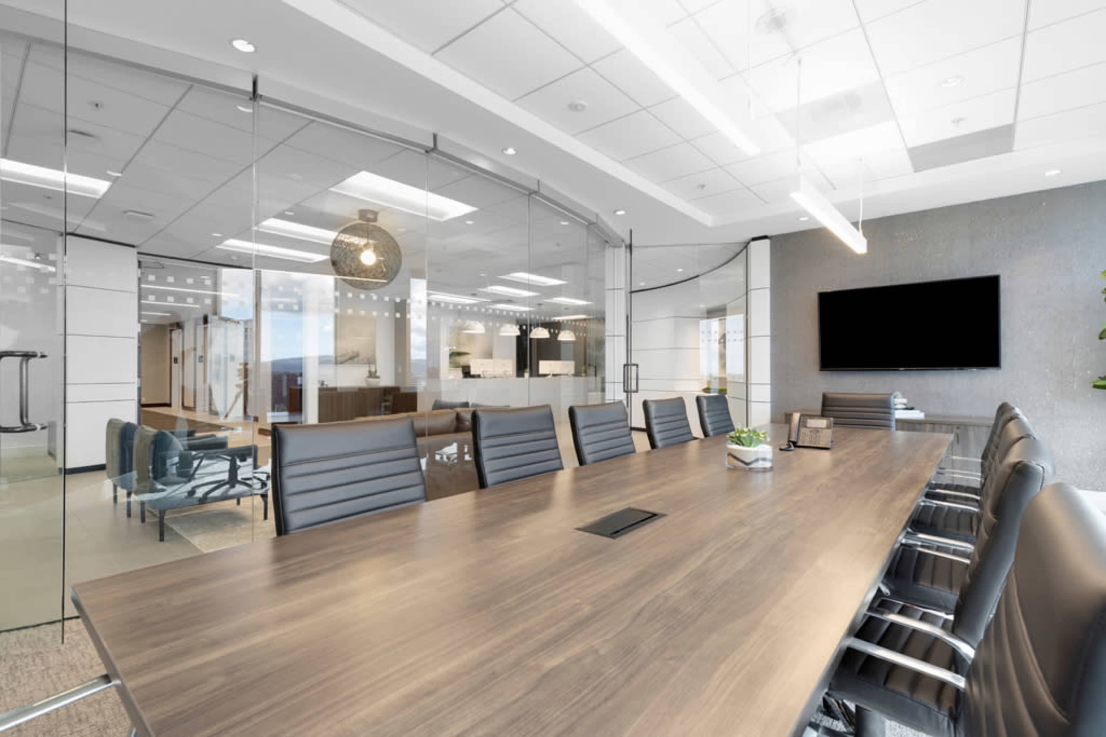 A modern conference room features a large wooden table surrounded by ergonomic chairs and a glass wall that separates it from the adjoining workspace.