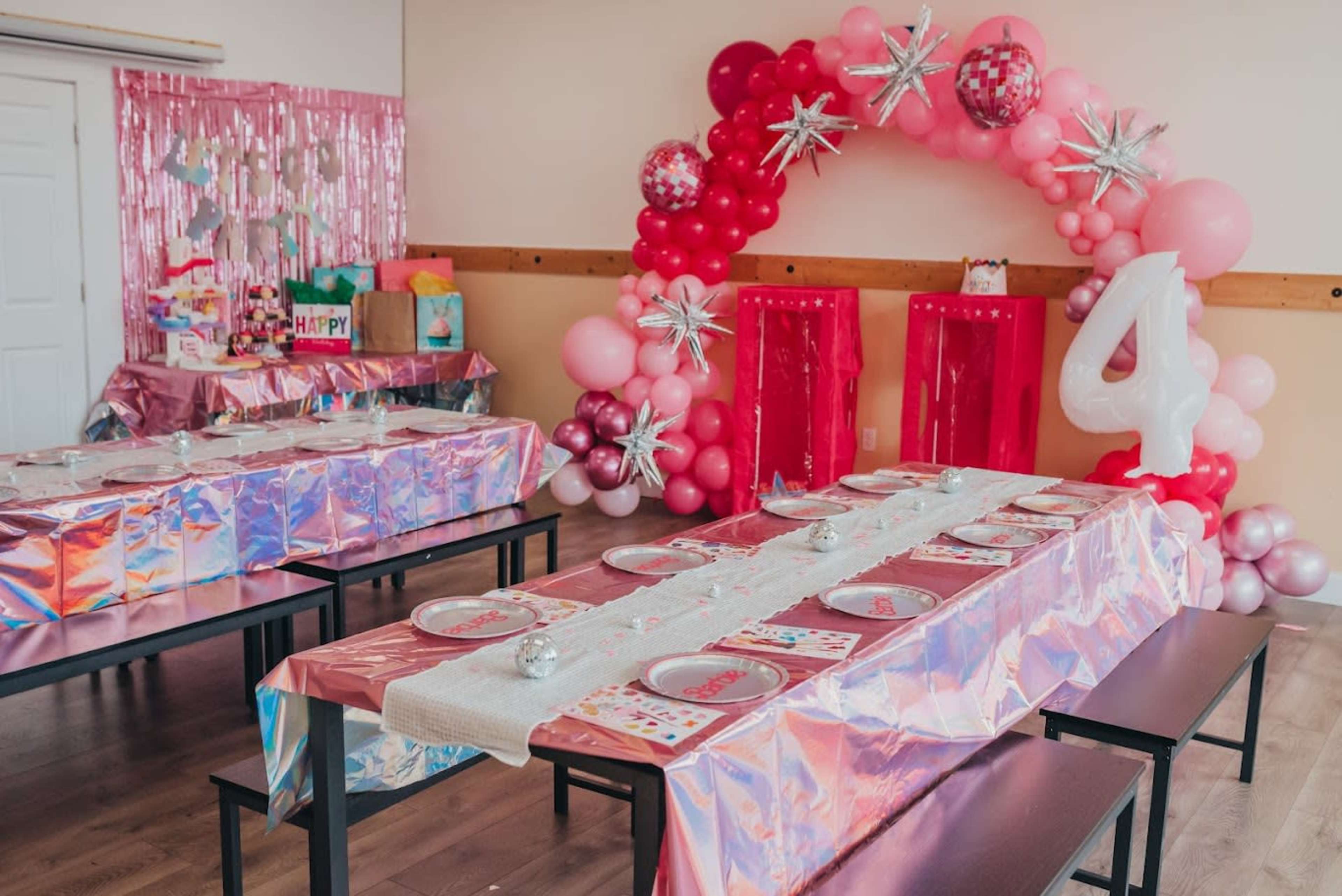 A party room is decorated with pink and silver elements, featuring a long table set with plates and a backdrop of balloons and a large number "4."