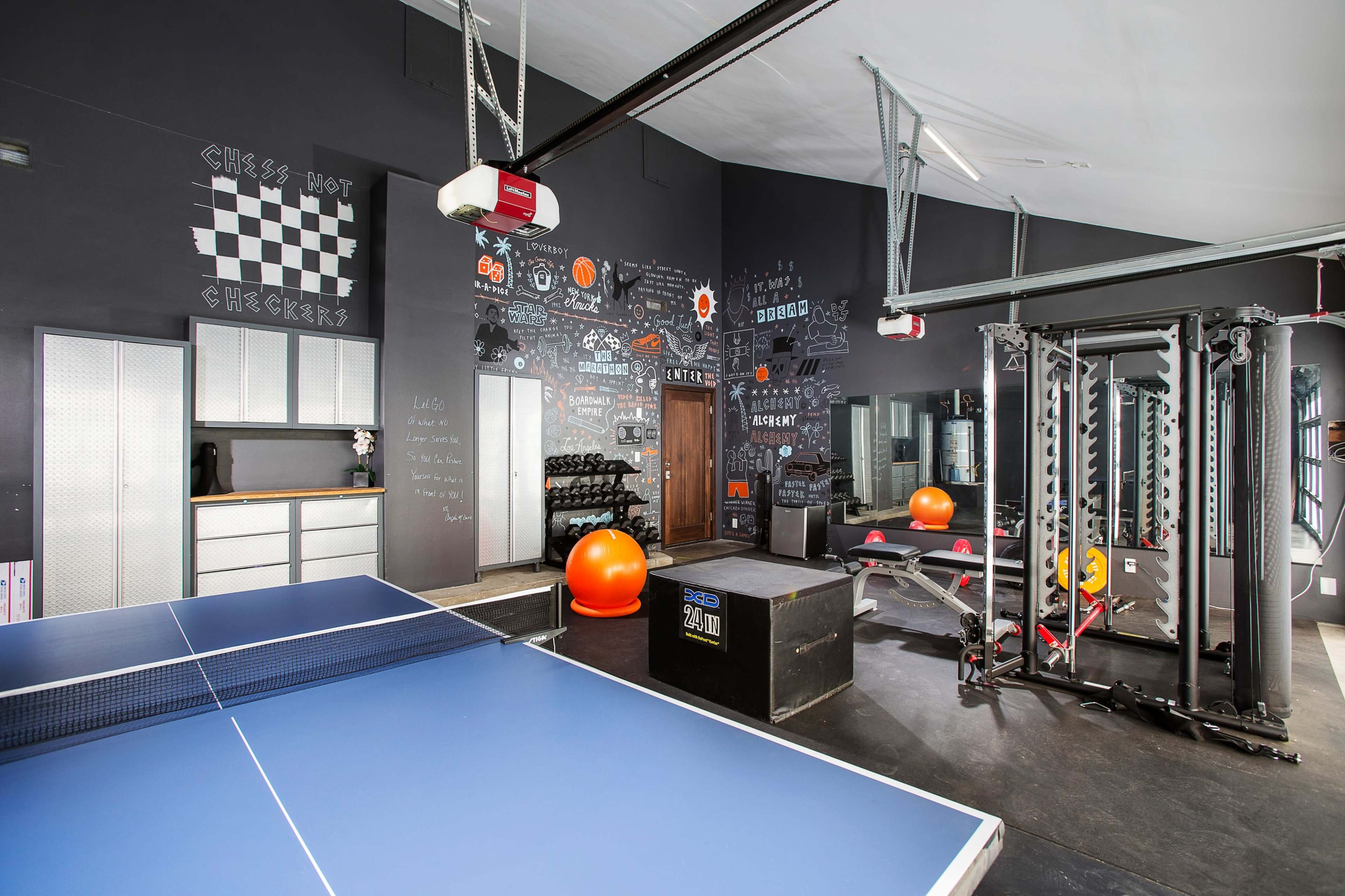 A modern gym interior featuring a ping pong table, a workout area with weights, and exercise equipment against a chalkboard wall adorned with various designs.